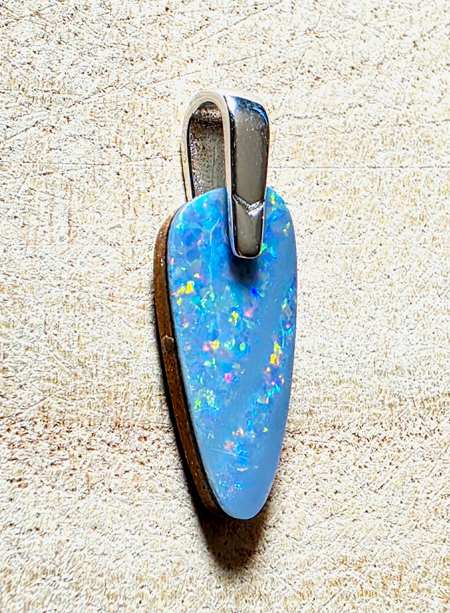 A teardrop-shaped blue opal pendant with a silver bail, displayed on a textured beige surface.