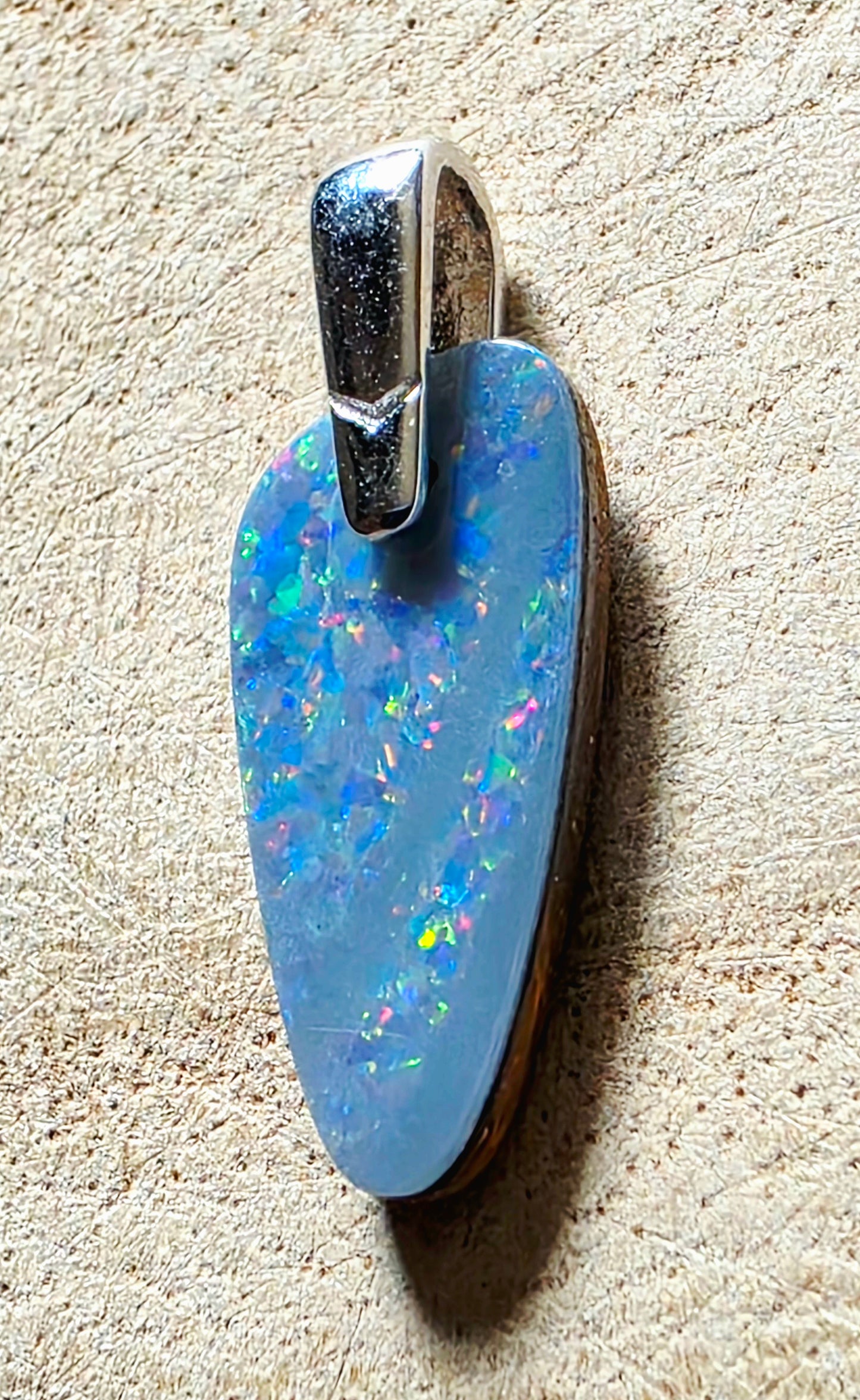 A teardrop-shaped blue opal pendant with a silver bail, resting on a textured beige surface.