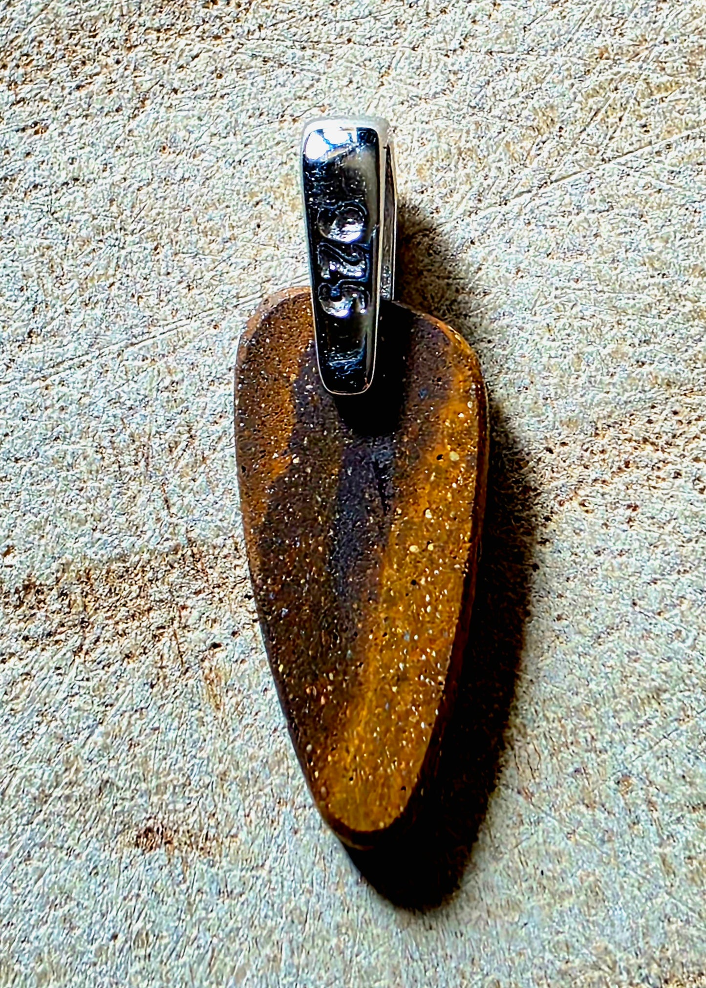 A polished brown stone pendant with tiny white flecks, attached to a silver bail shown on a textured beige surface.