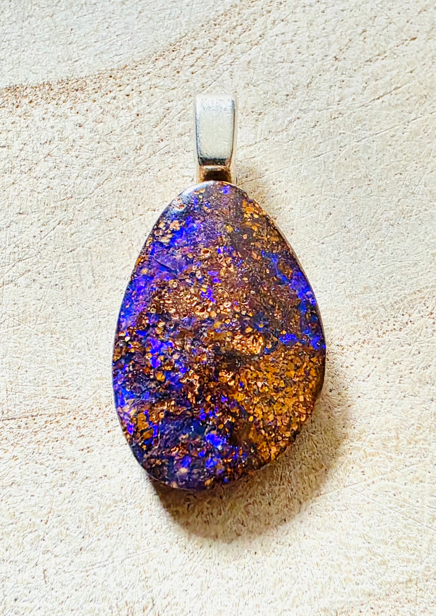 A teardrop-shaped opal pendant with vibrant blue, purple, and orange play-of-colour, set in a silver-toned bail, displayed on a textured beige surface.