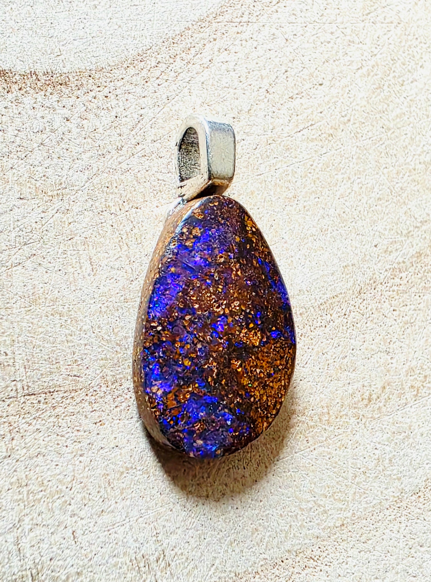 A teardrop-shaped opal pendant with vibrant blue and orange play-of-colour, set in a silver-toned metal bezel, displayed on a textured beige surface.