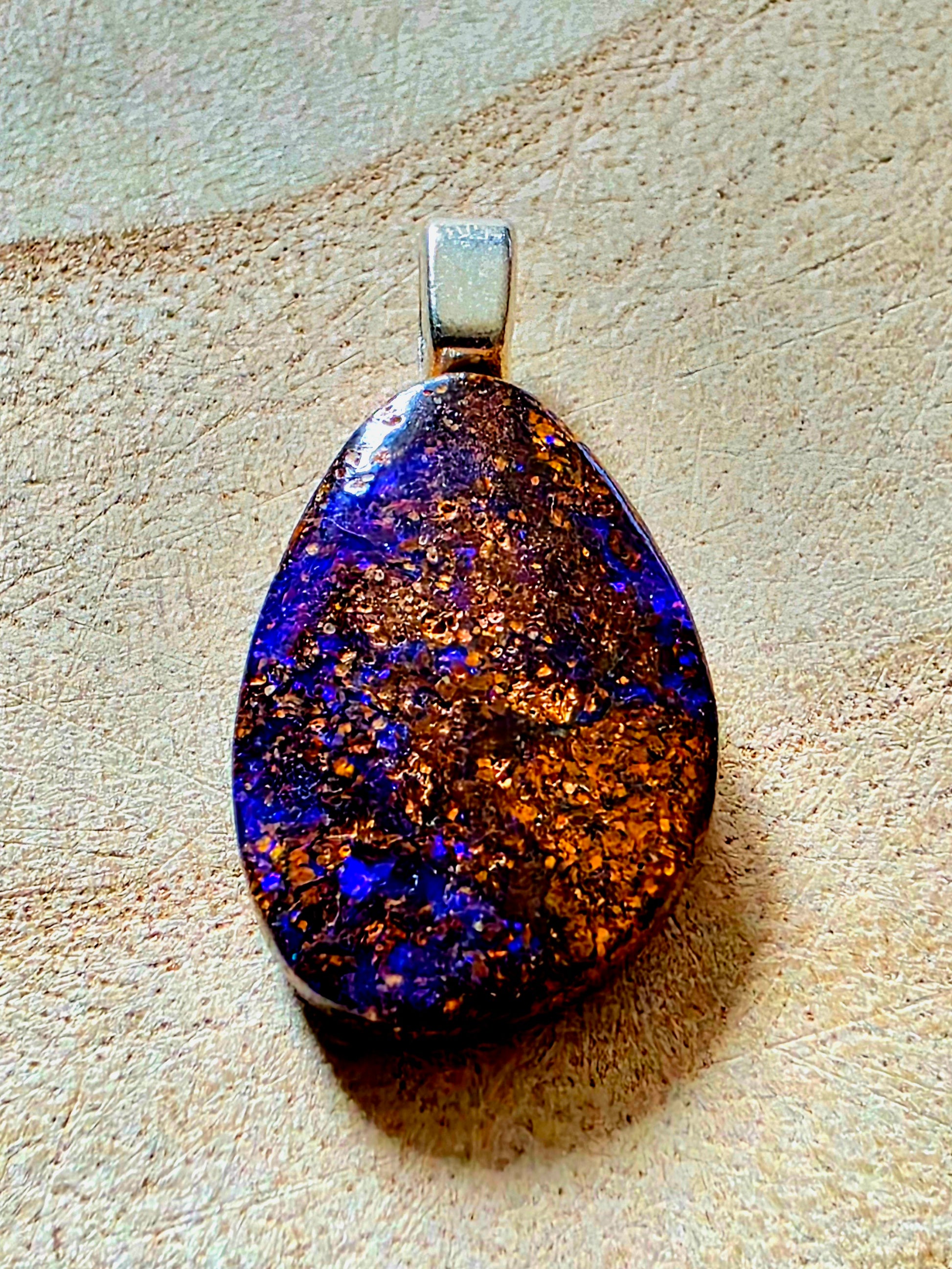 A teardrop-shaped pendant featuring a highly iridescent opal with shimmering blue, purple, and metallic gold patterns, set in a silver-tone bail, resting on a textured beige surface.