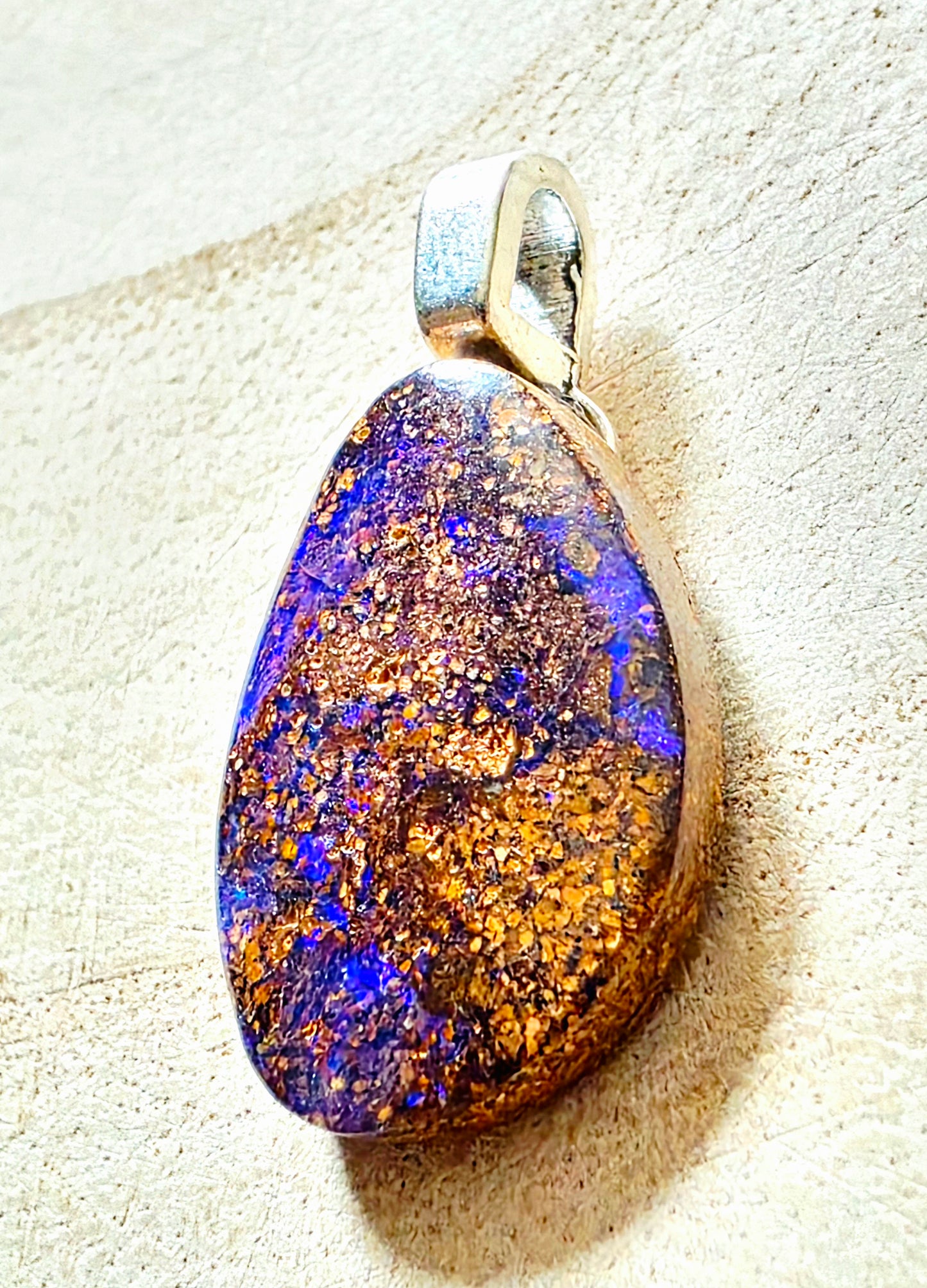 A teardrop-shaped pendant featuring a highly iridescent opal stone with shimmering gold and deep blue-purple hues, set in a polished silver-tone metal bezel with a bail, resting on a textured beige surface.