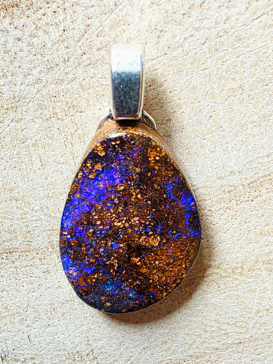 A teardrop-shaped pendant featuring a natural iridescent opal with vibrant blue, purple, and gold flashes, set in a silver-toned metal bezel on a textured beige background.