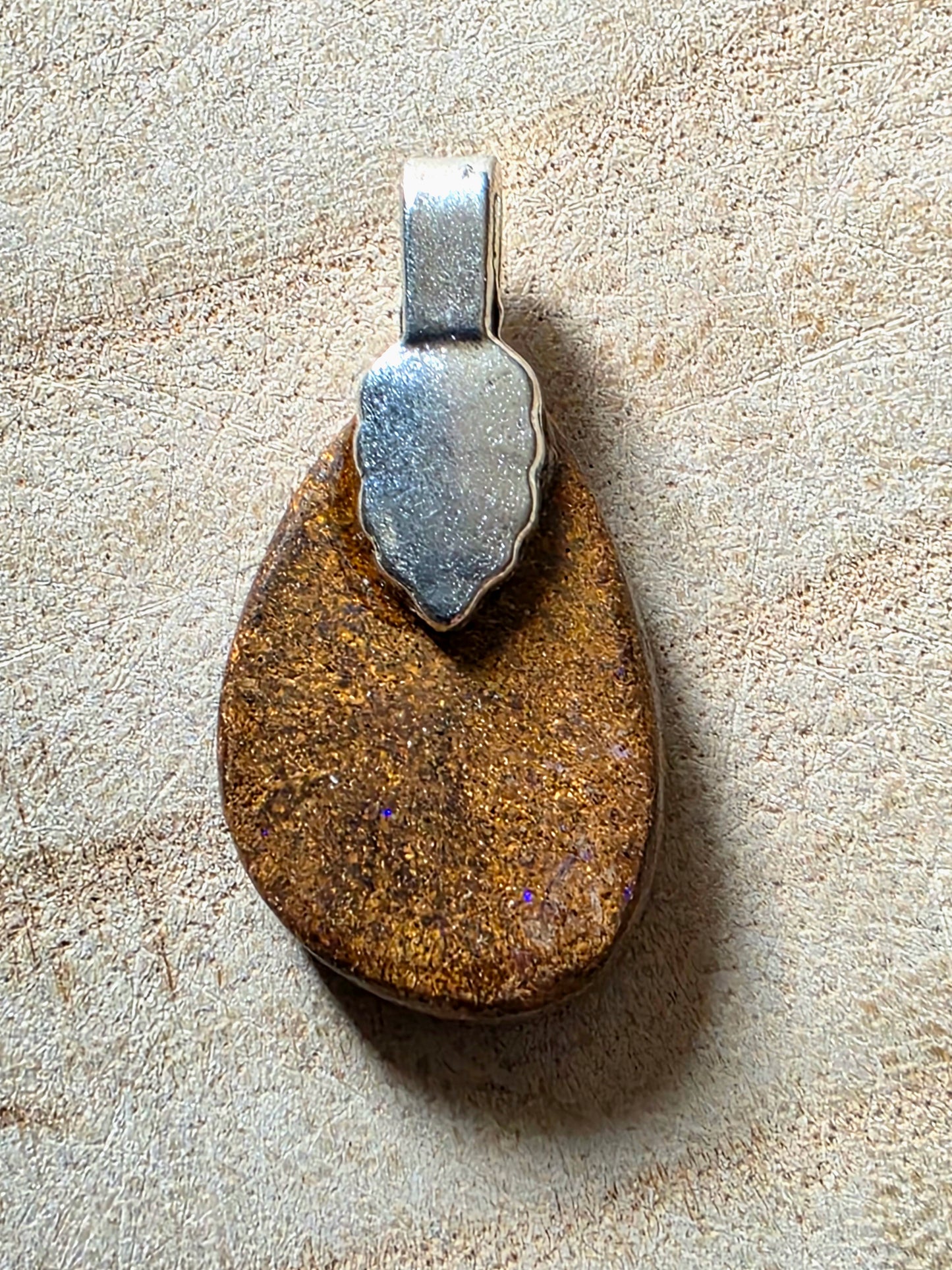 A teardrop-shaped pendant featuring a polished brown and gold opal stone set in a silver-toned metal bail, displayed on a textured beige surface.