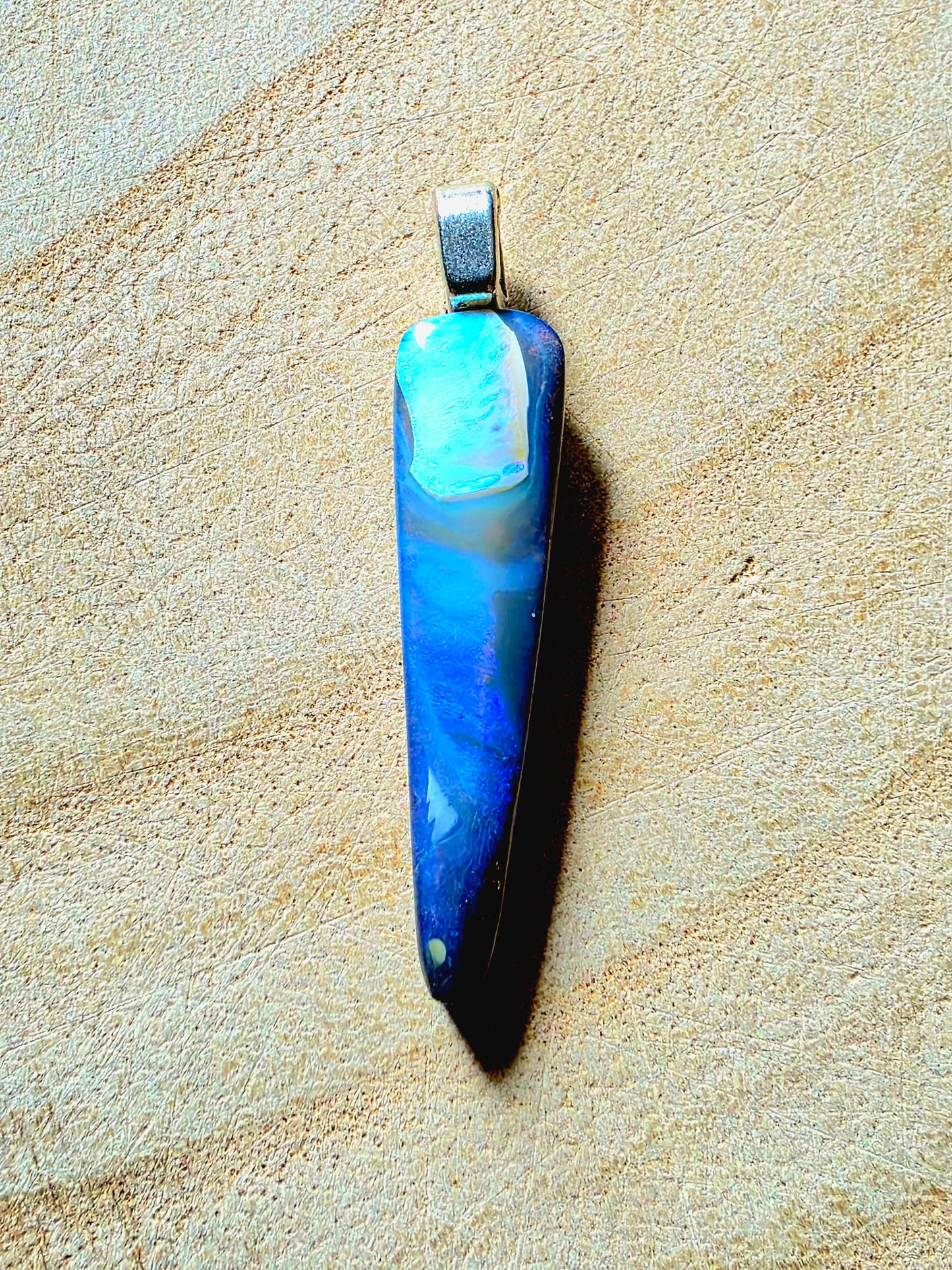 A vertical teardrop-shaped pendant made of blue and white marbled opal, set in a silver-toned metal bail, resting on a textured beige surface.