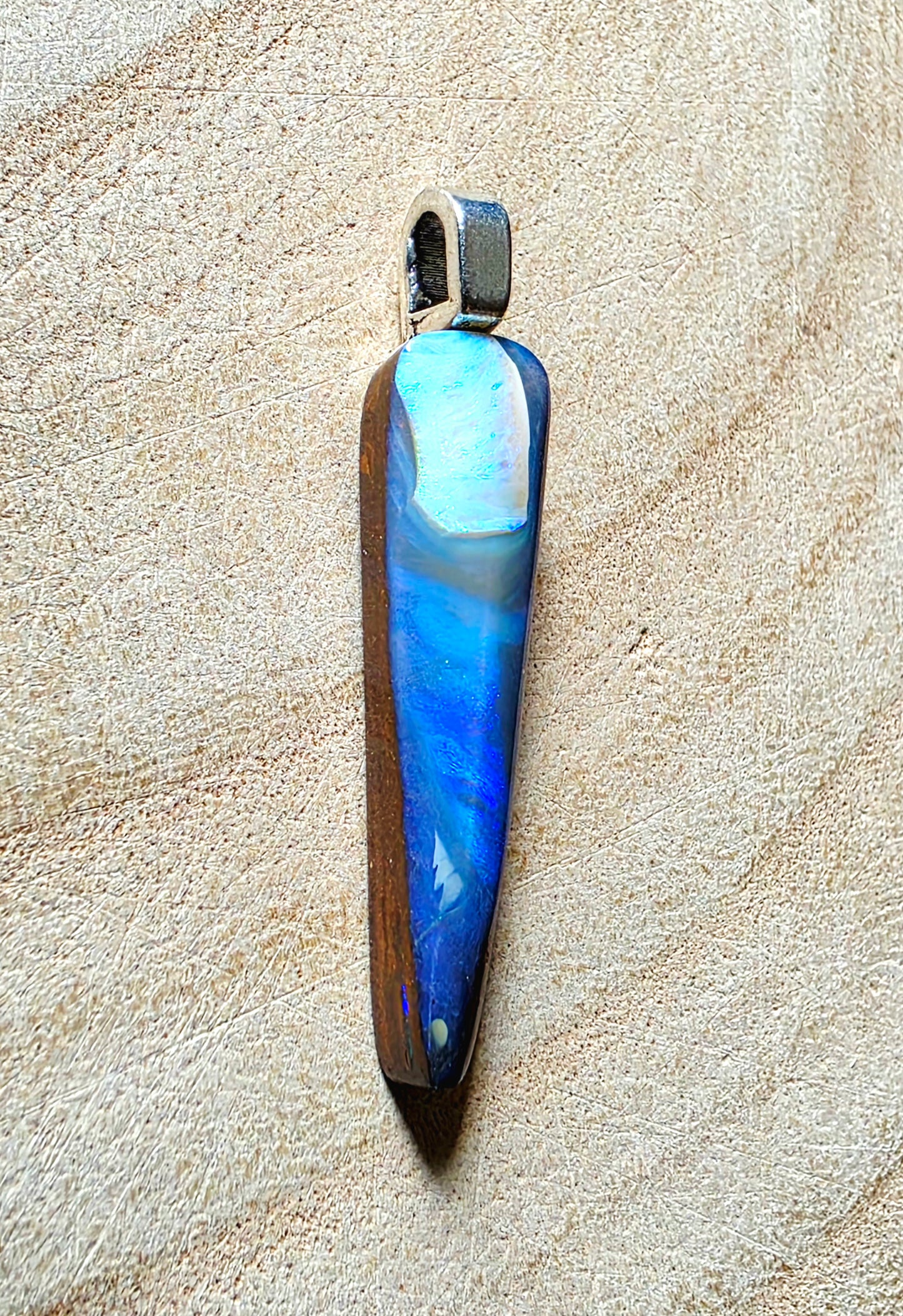 A teardrop-shaped pendant featuring a marbled blue and white opal with a dark brown wood-grain side, suspended from a silver-toned metal loop, displayed on a textured beige surface.