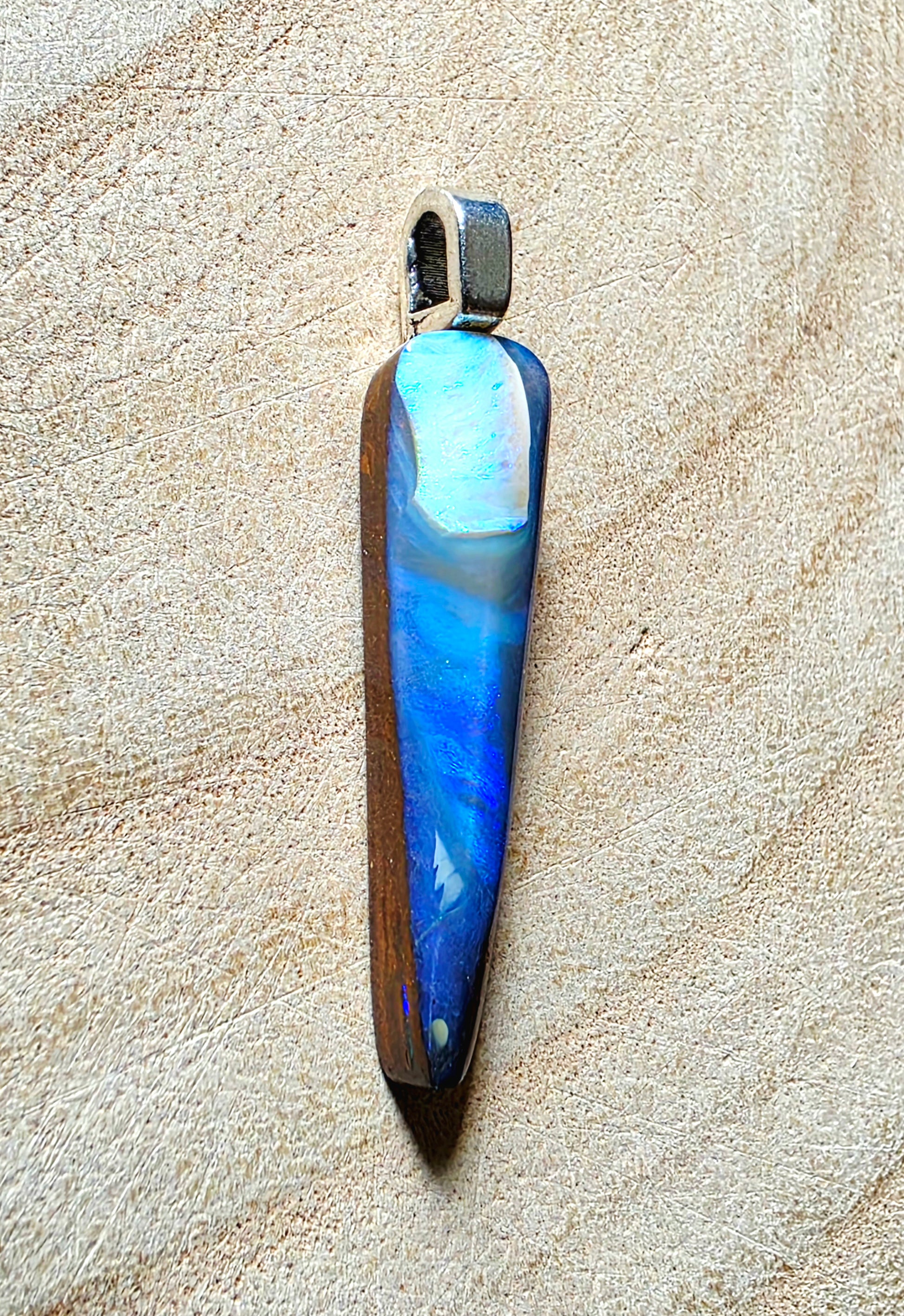 A teardrop-shaped pendant featuring a marbled blue and white opal with a dark brown wood-grain side, suspended from a silver-toned metal loop, displayed on a textured beige surface.