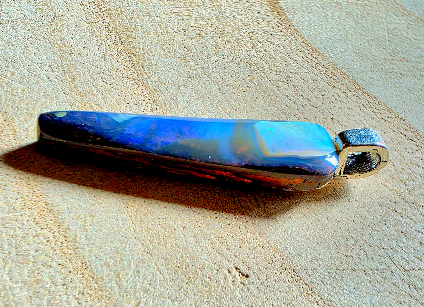 A polished iridescent opal pendant with blue, purple, and gold colour shifts, set in a silver-toned bezel with a bail, resting on a textured beige surface.