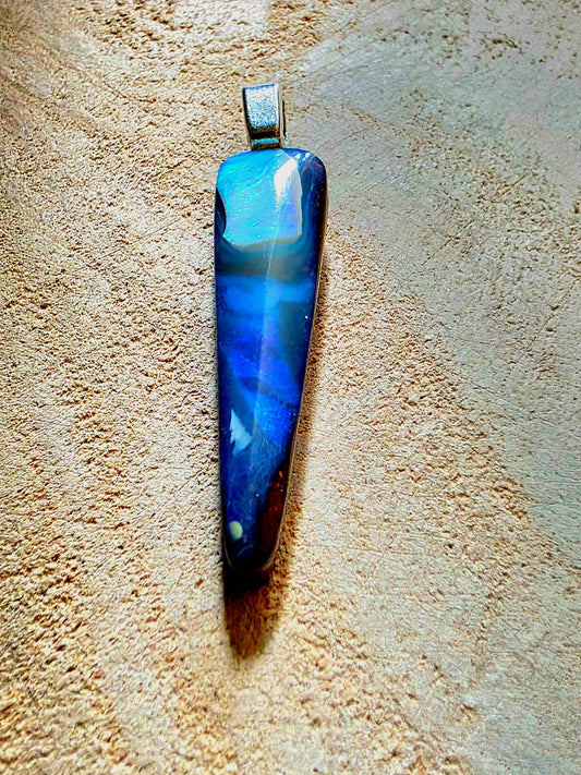 A blue and white marbled opal pendant with a silver bail, displayed vertically on a textured beige stone surface.
