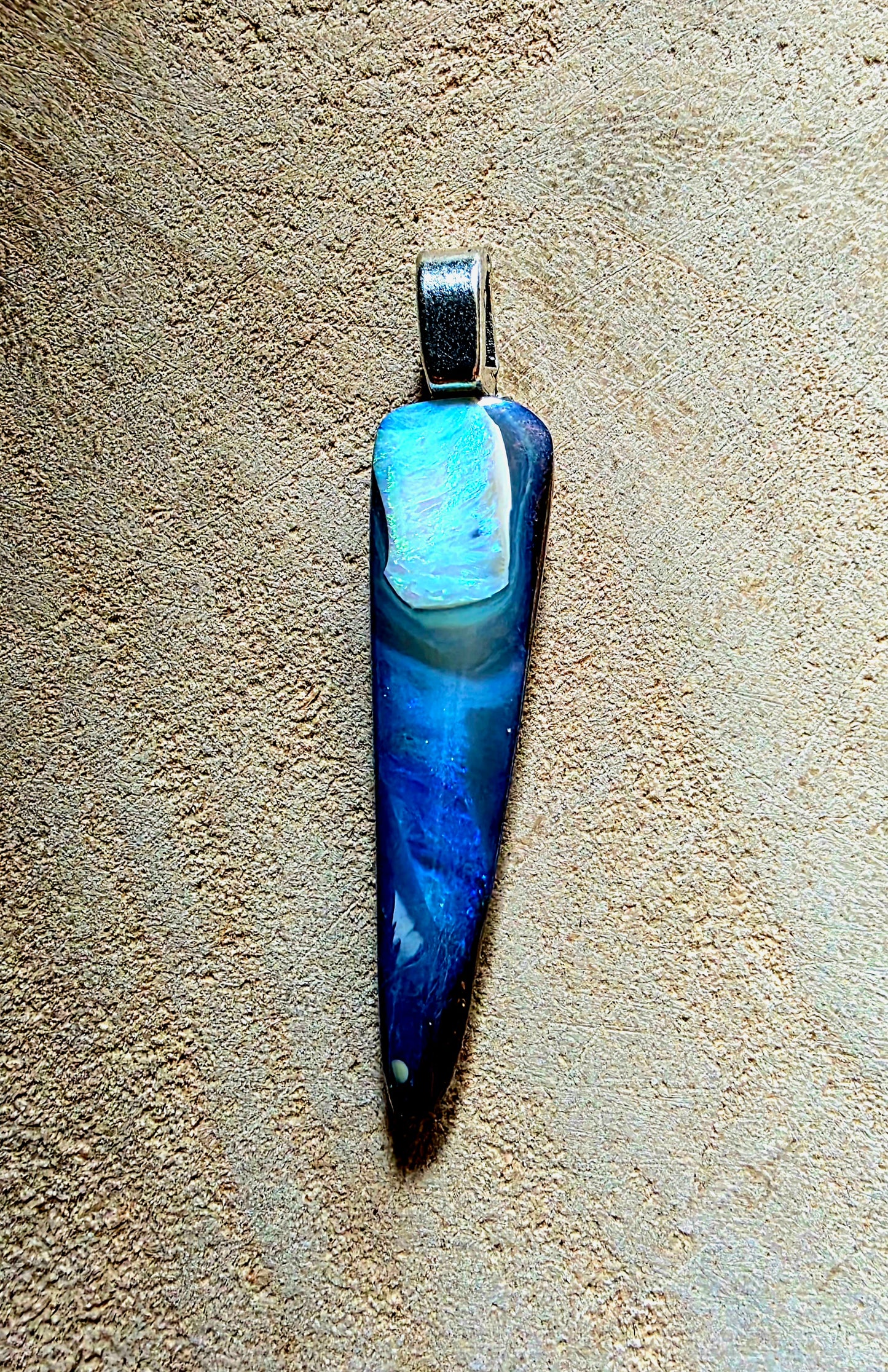 A teardrop-shaped pendant featuring a marbled blue and white opal stone body with a silver-toned bail, displayed against a textured beige background.