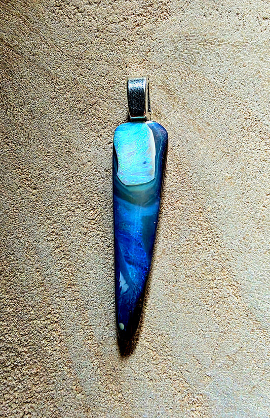 A teardrop-shaped pendant featuring a marbled blue and white opal stone body with a silver-toned bail, displayed against a textured beige background.