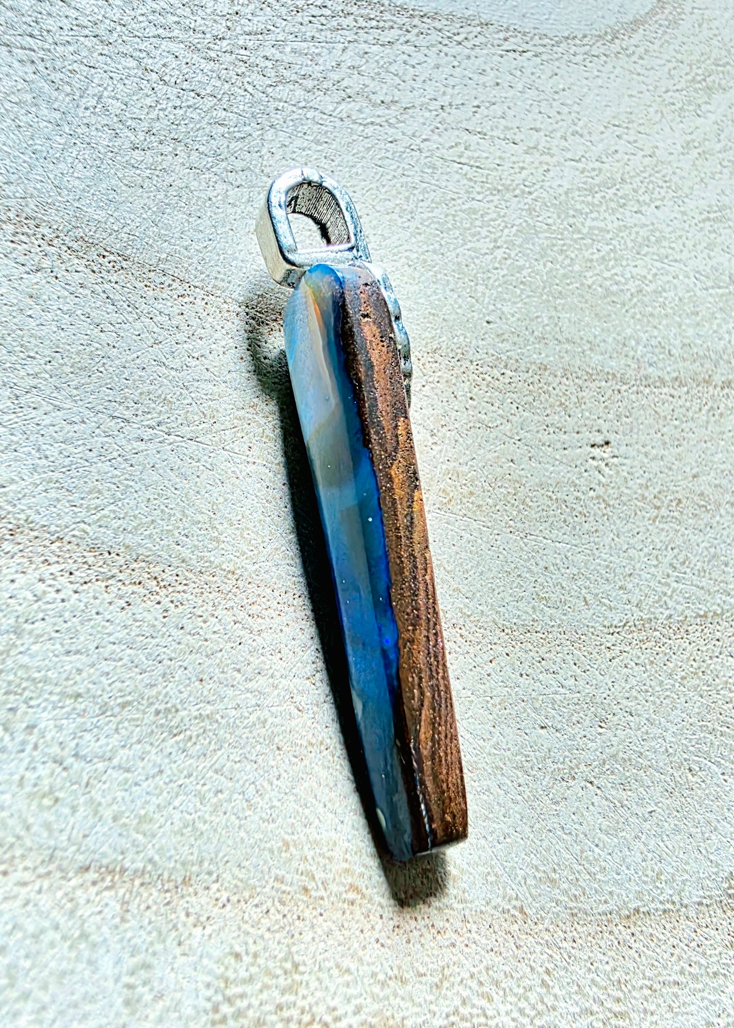 A handmade pendant featuring a polished opal slice with natural brown grain and a blue resin inlay, attached to a silver metal loop, displayed on a textured beige surface.