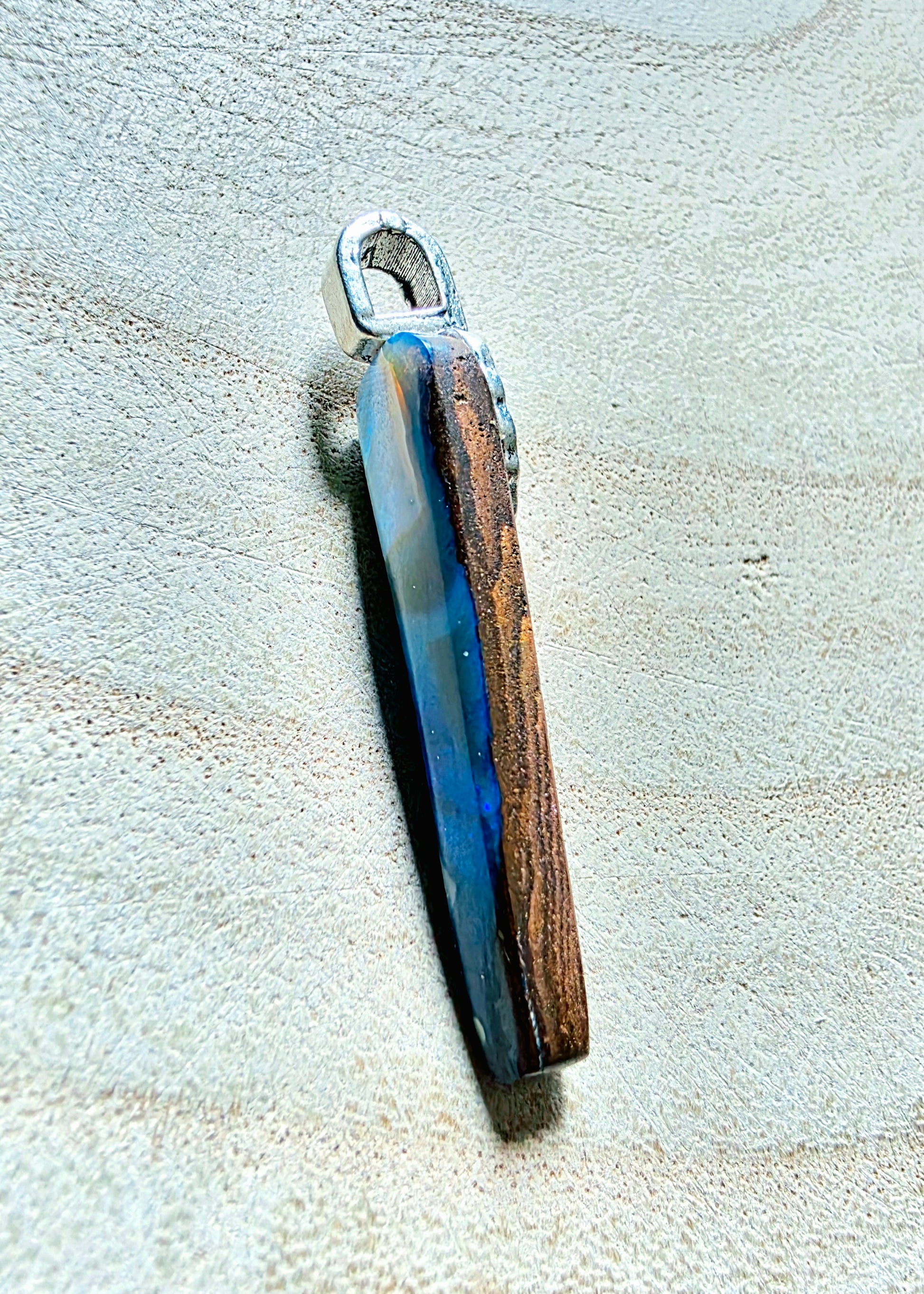 A handmade pendant featuring a polished opal slice with natural brown grain and a blue resin inlay, attached to a silver metal loop, displayed on a textured beige surface.