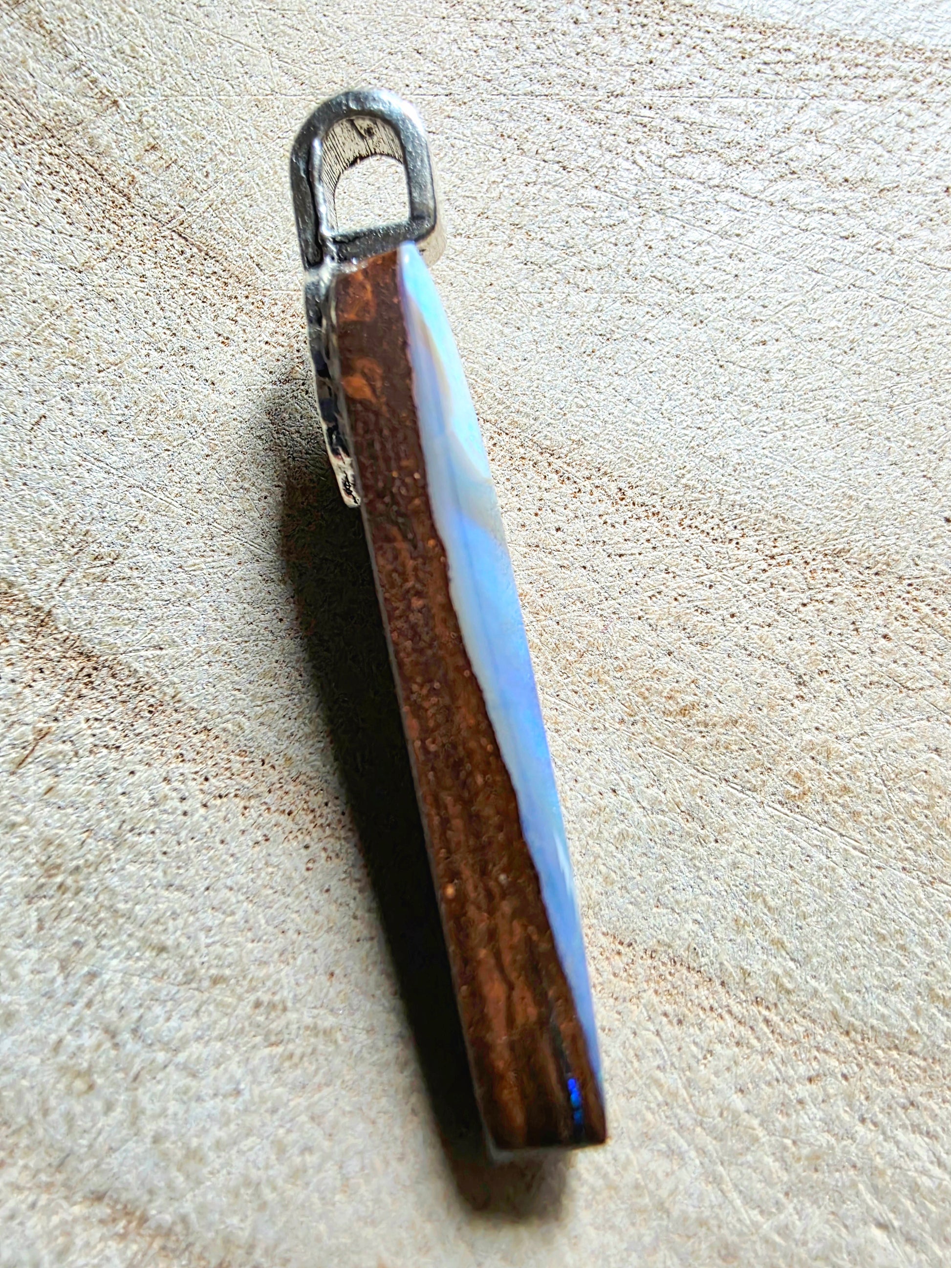 A rectangular pendant with a silver-tone metal loop at the top, featuring a natural brown wood grain center and a light blue iridescent side, resting on a textured beige surface.