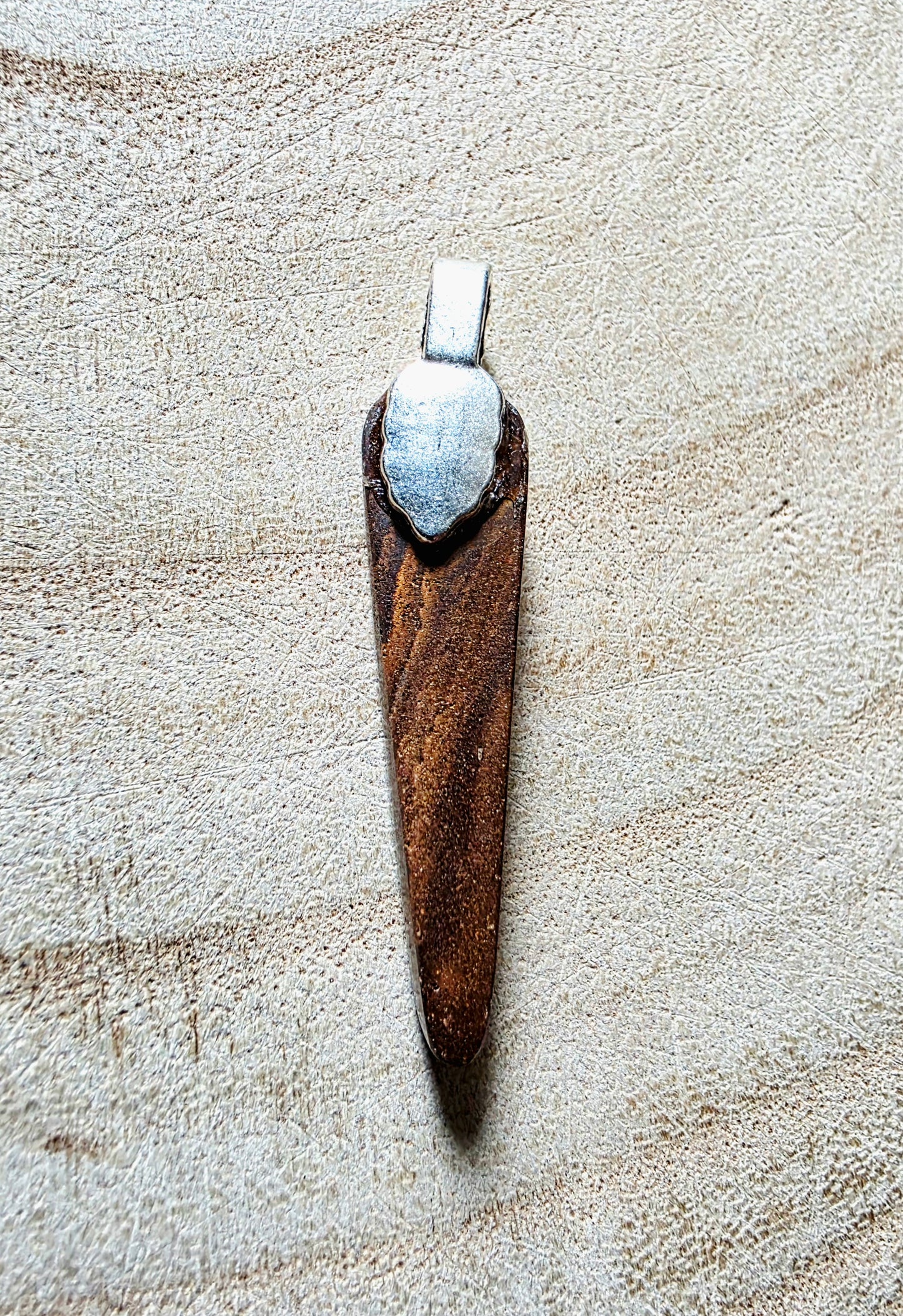 A handmade opal and metal pendant lies on a textured beige fabric background. The pendant features a dark brown body with a silver-toned metal cap at the top.