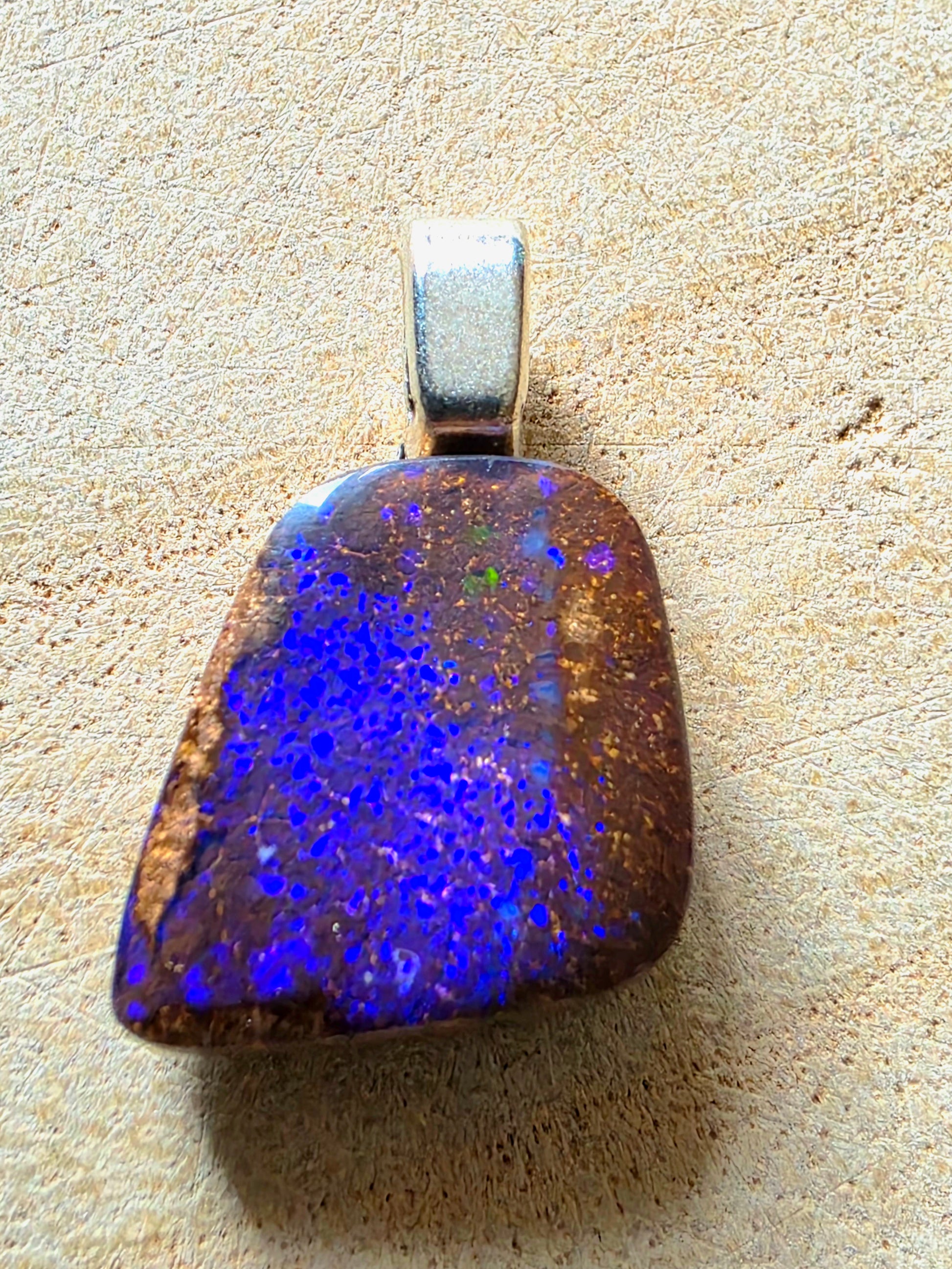 A natural opal pendant with a silver bail, displayed on a textured beige surface. The opal features a rich color play of vibrant blue, purple, and green flashes over a dark brown and gold matrix.