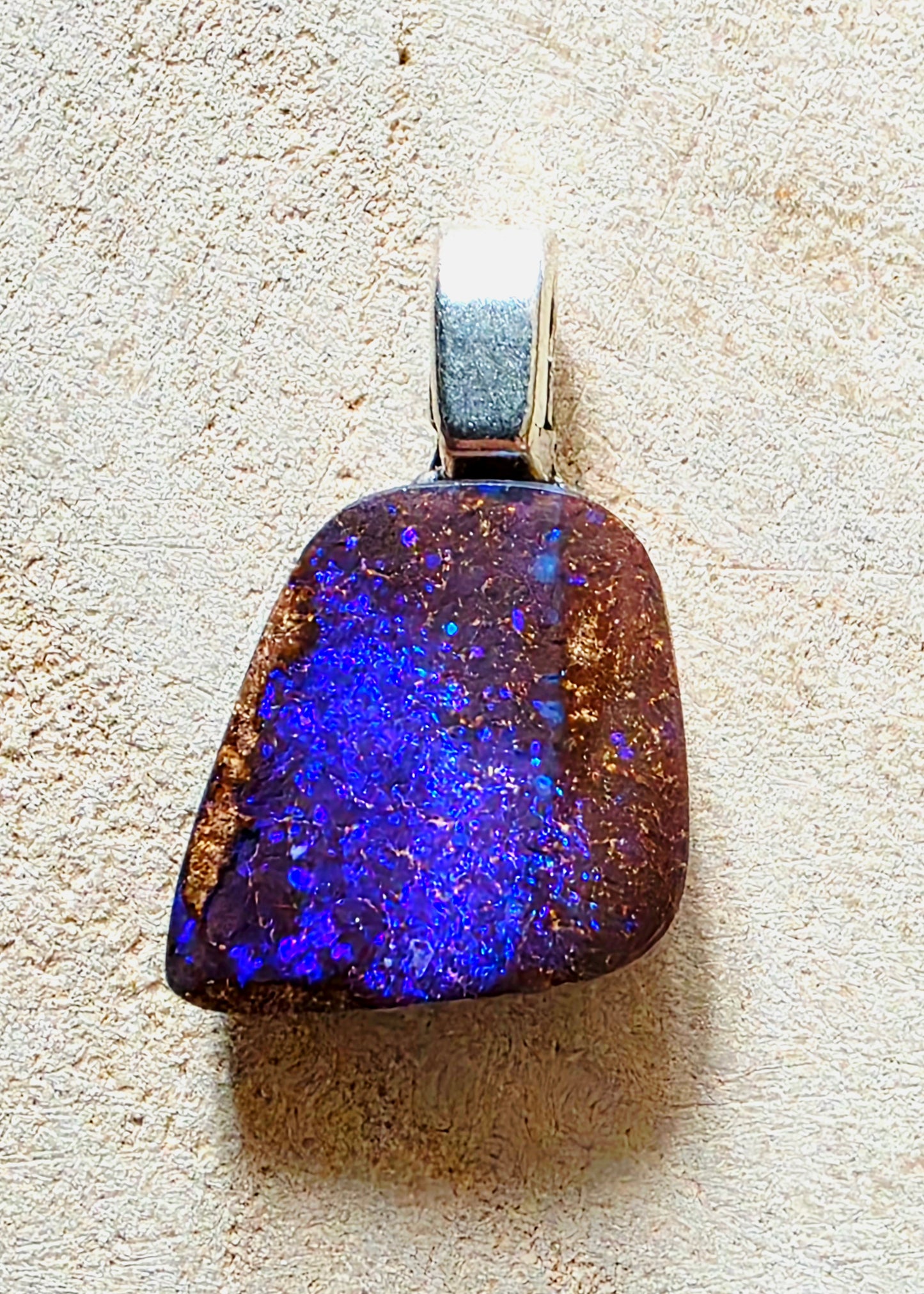 A natural blue opal pendant with a silver bail, displayed on a textured beige surface.