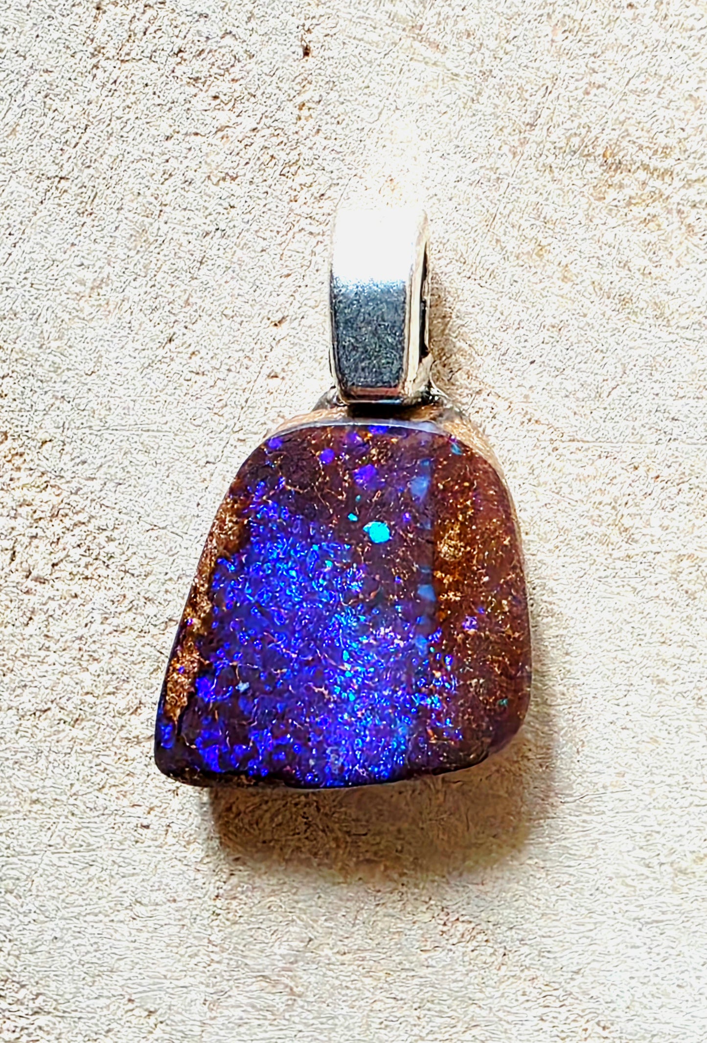 A raw opal pendant with a silver bail, resting on a textured beige surface. The opal displays a rich color play with vibrant blue, purple, and orange flashes over a dark brown base.