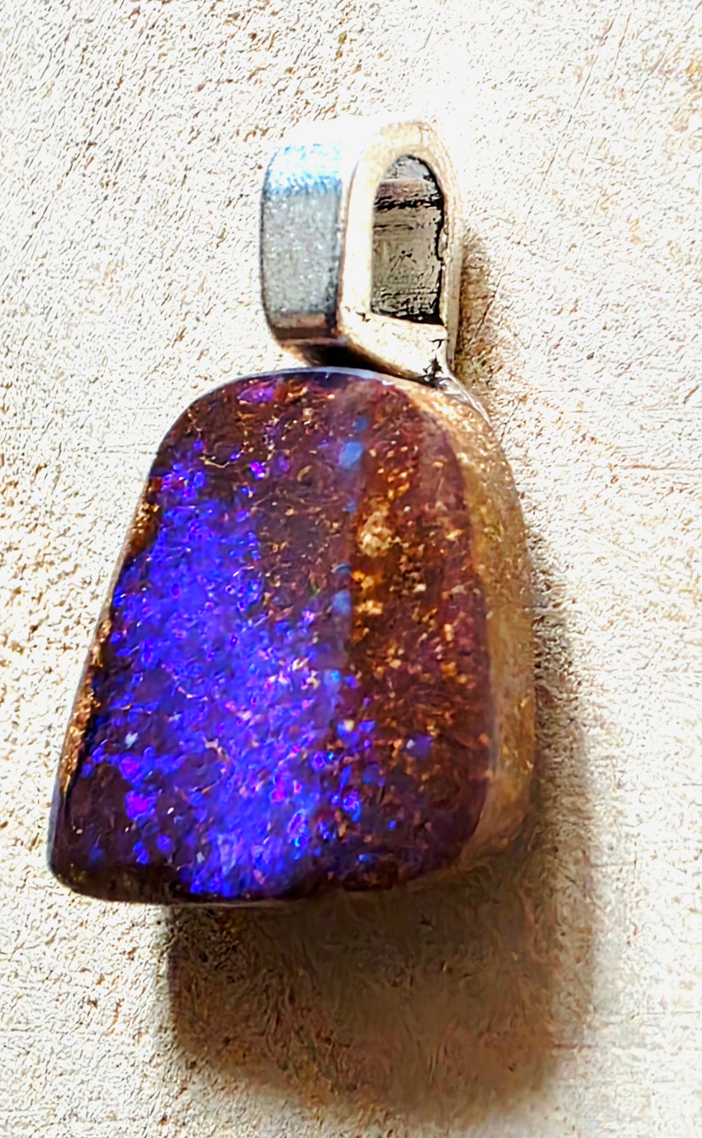 A natural teardrop-shaped opal pendant with a silver bail, displayed on a textured beige surface. The opal exhibits a vibrant play-of-color with bright blue, purple, and brown iridescent flashes.