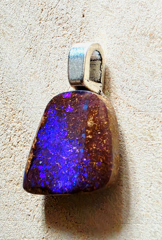 A teardrop-shaped pendant featuring a vibrant blue and purple cosmic glitter stone with gold flecks, set in a silver-toned metal bezel, shown on a textured beige background.