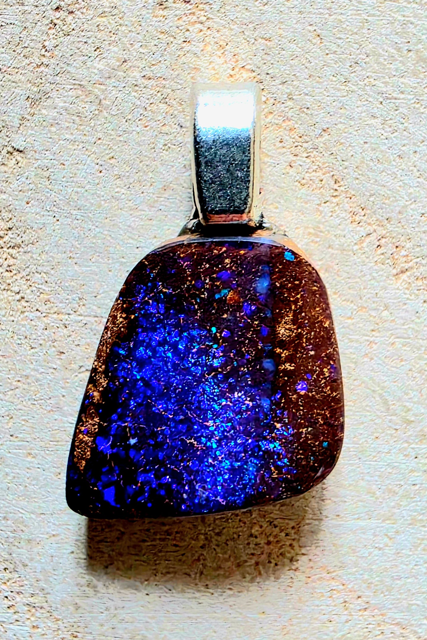 A teardrop-shaped pendant featuring a vibrant blue and purple cosmic glitter stone with gold flecks, set in a silver-toned metal bezel, shown on a textured beige background.