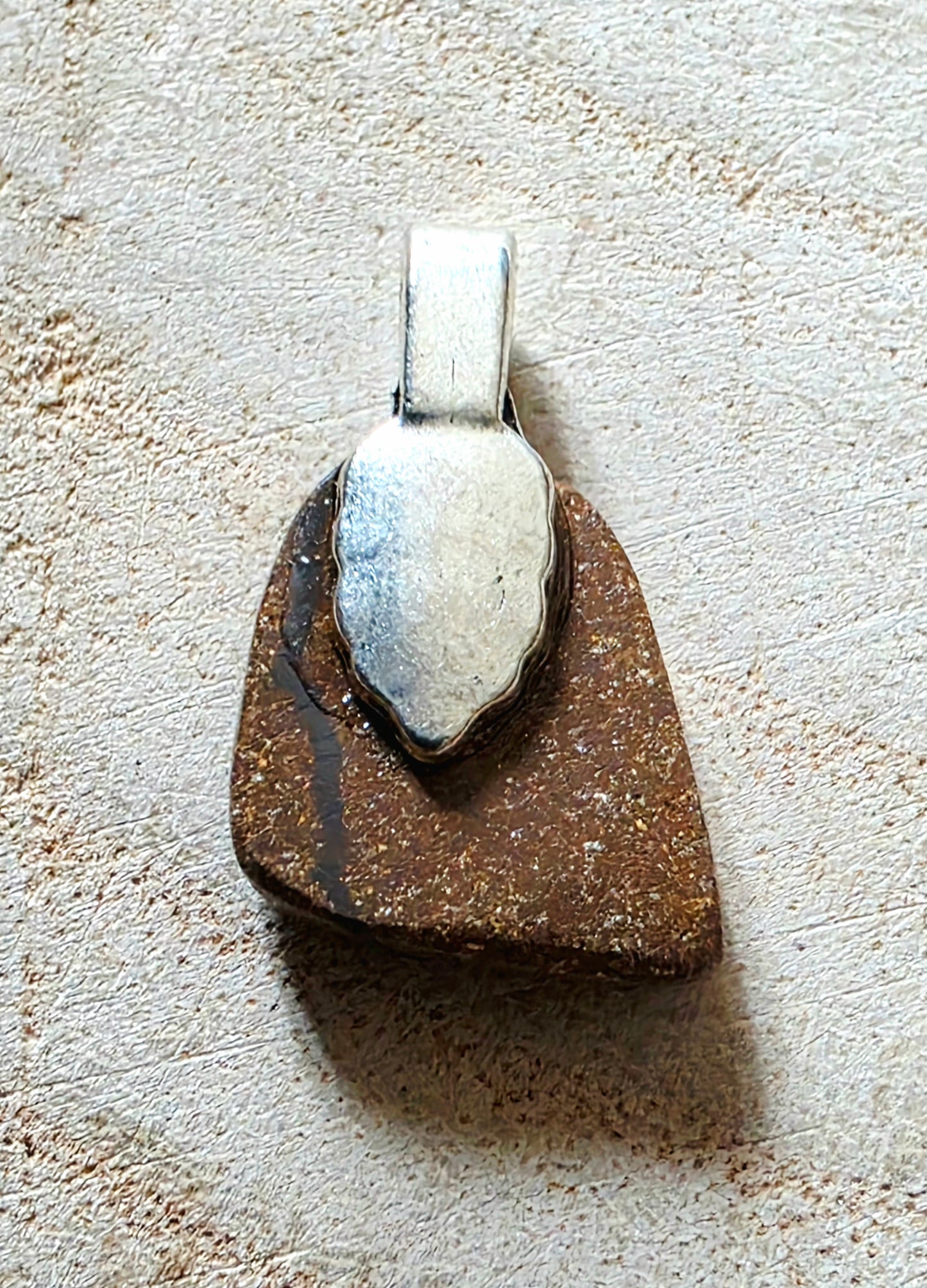 A handmade pendant featuring a rustic brown stone base with a polished silver leaf-shaped accent on top, resting on a textured beige surface.