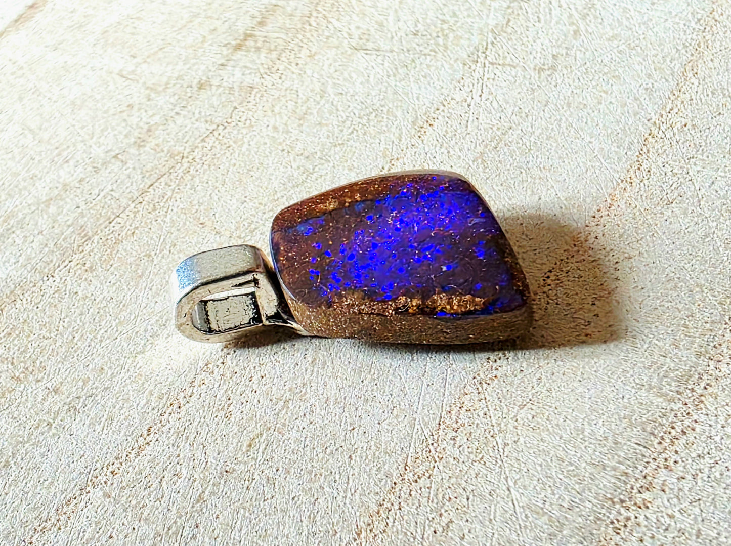 A square pendant featuring a natural blue opal stone with a brown matrix, attached to a silver-toned metal bail, displayed on a textured beige surface.
