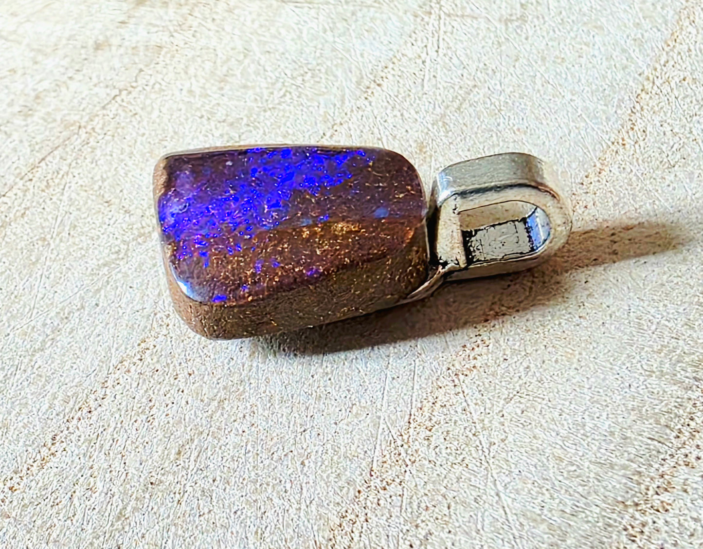 A polished pendant featuring a rectangular brown opal stone with vibrant blue and purple iridescent flashes, set in a silver bezel, resting on a textured beige surface.