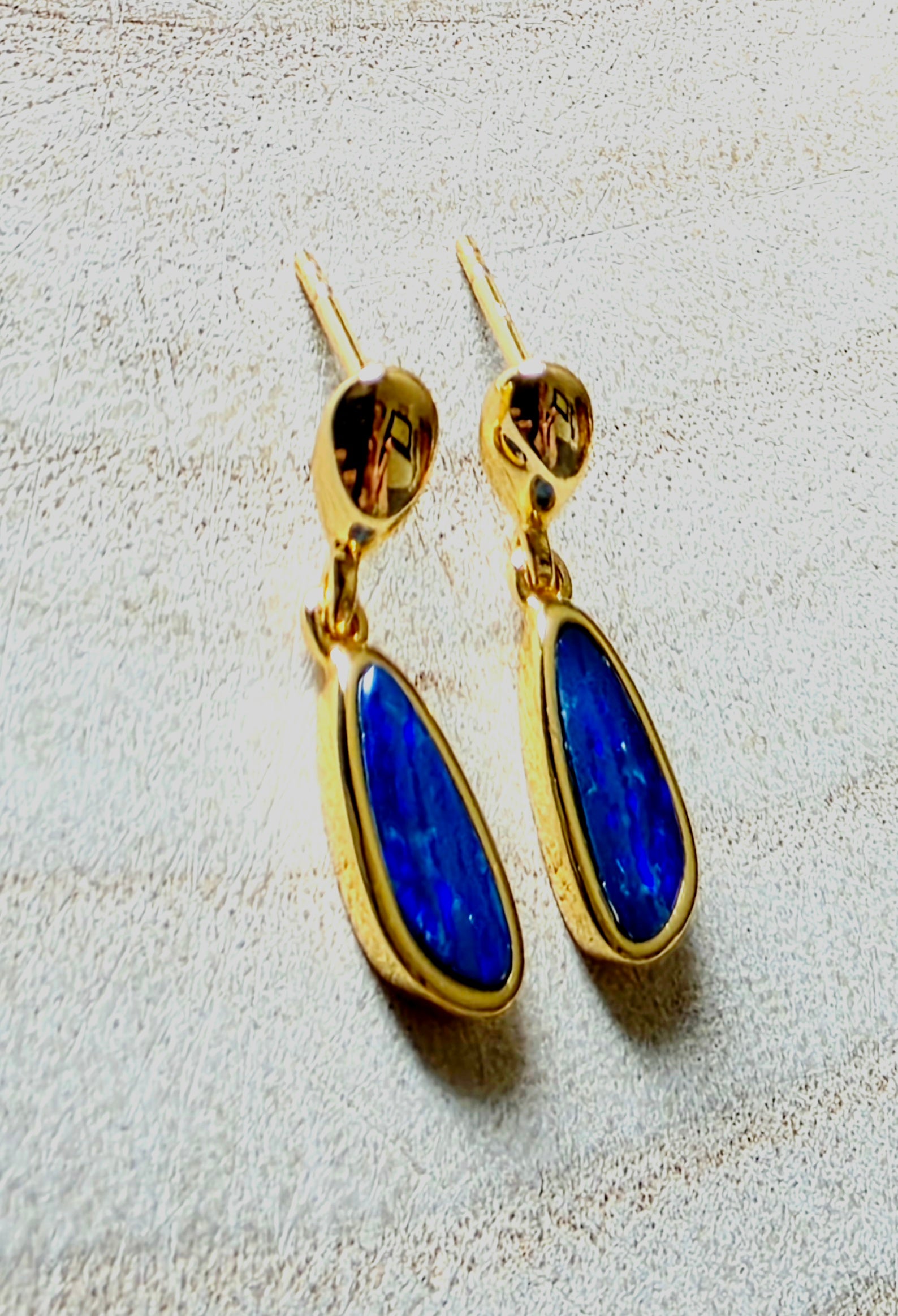 A pair of gold drop earrings with teardrop-shaped deep blue opals, displayed on a textured light beige surface.