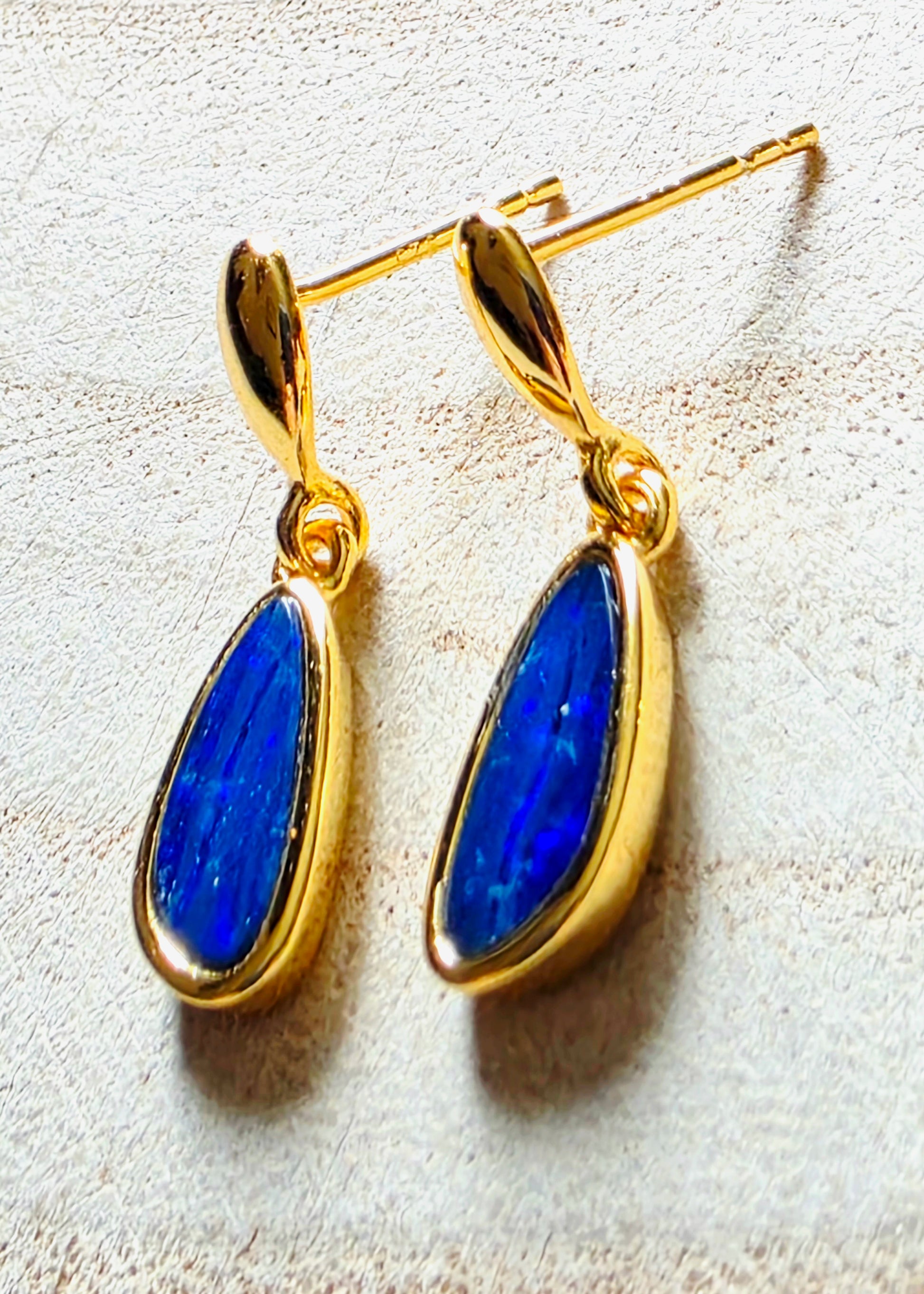 A pair of gold drop earrings with teardrop-shaped blue opal gemstone pendants, displayed on a textured light beige surface.