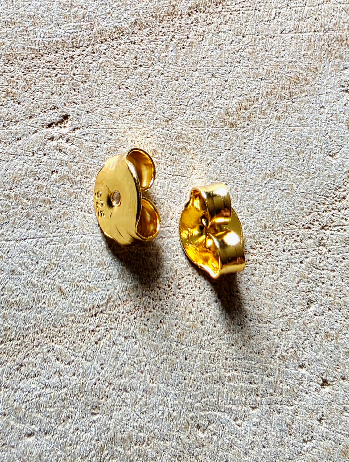 A pair of gold-toned earrings shown from the back on a textured beige surface, displaying the flat back and post details.