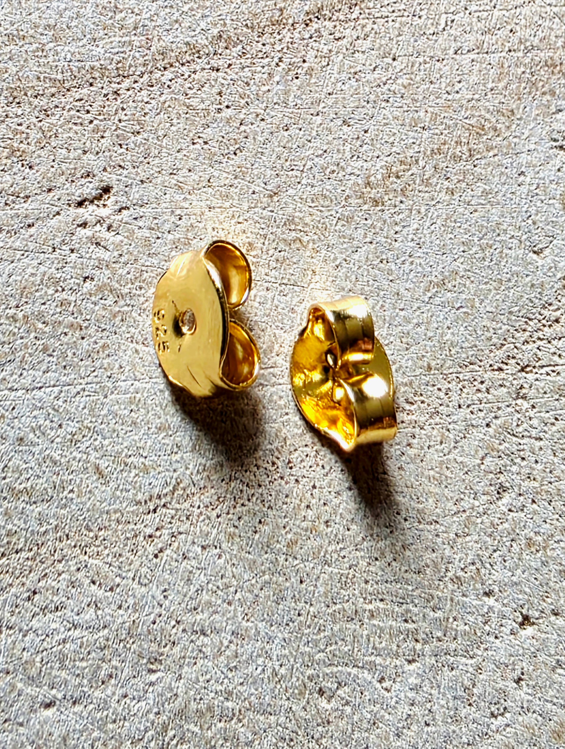 A pair of gold-toned earrings shown from the back on a textured beige surface, displaying the flat back and post details.