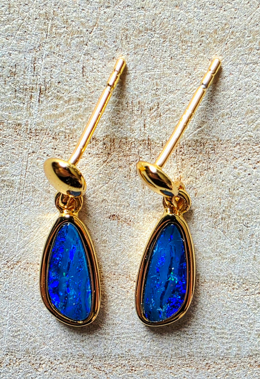 A pair of gold drop earrings with teardrop-shaped blue opal stones, displayed on a textured beige surface.