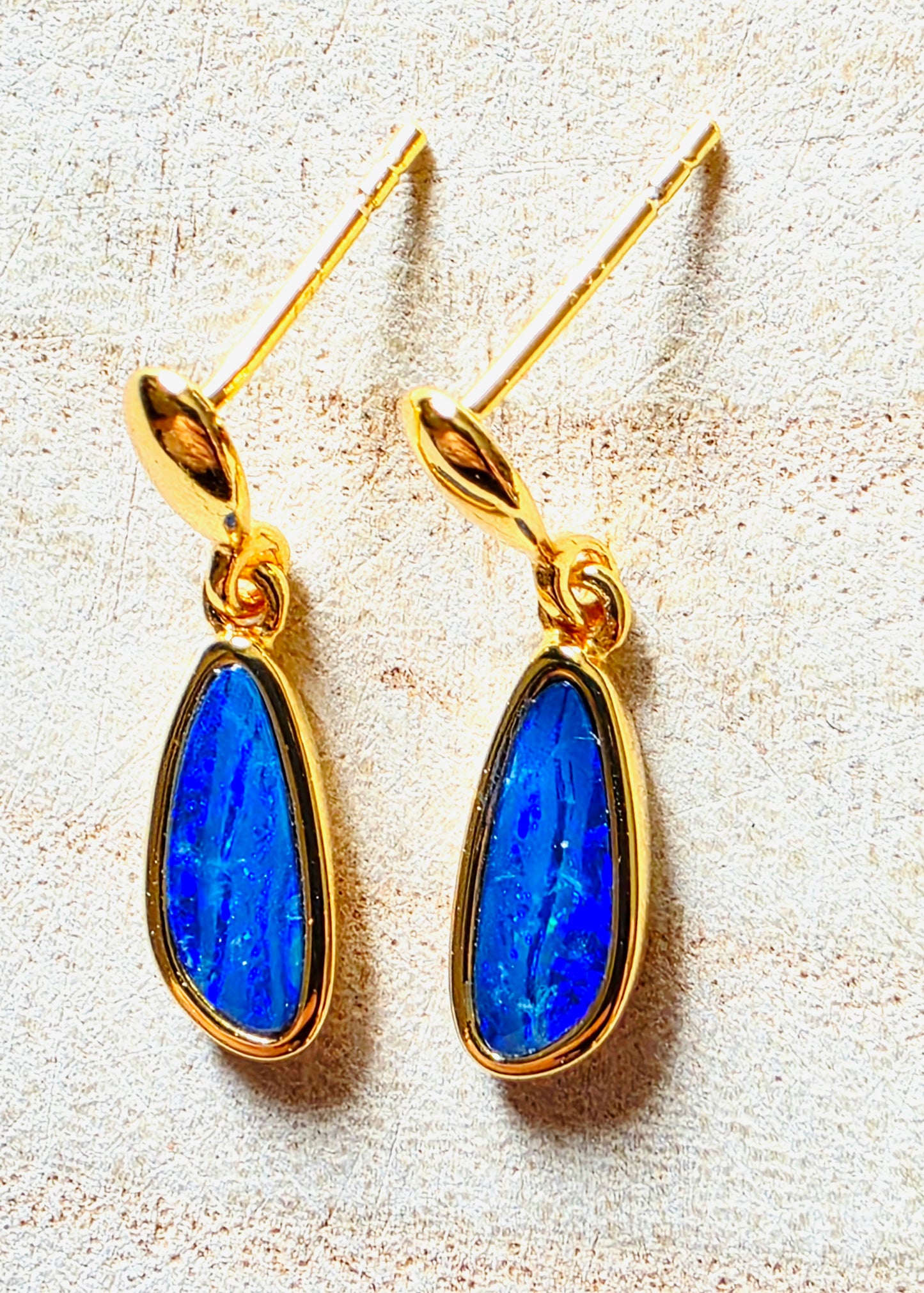 A pair of gold drop earrings with teardrop-shaped blue opal gemstones, displayed on a textured beige surface.
