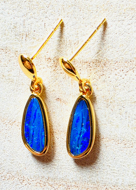 A pair of gold drop earrings with teardrop-shaped blue opal gemstones, displayed on a textured beige surface.