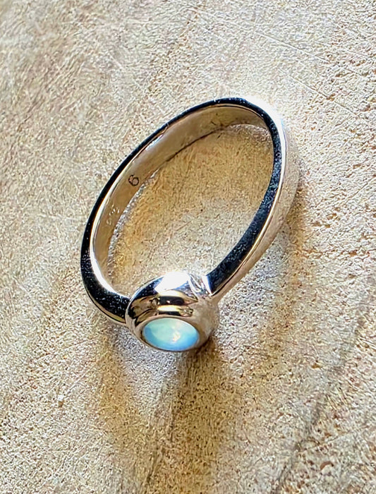 A sterling silver ring with a smooth, polished band and a bezel-set oval cabochon stone showing a soft blue-green iridescent luster, resting on a textured beige surface.