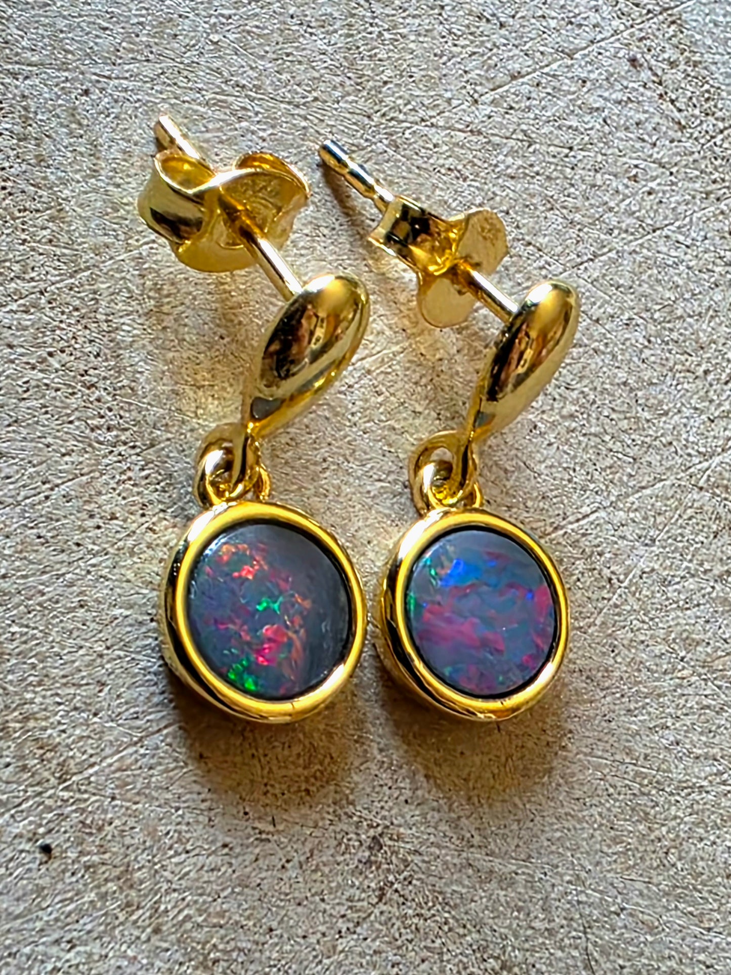 A pair of gold drop earrings with round bezel-set black opal gemstones showing vibrant blue, green, and pink flashes, displayed on a textured beige surface.