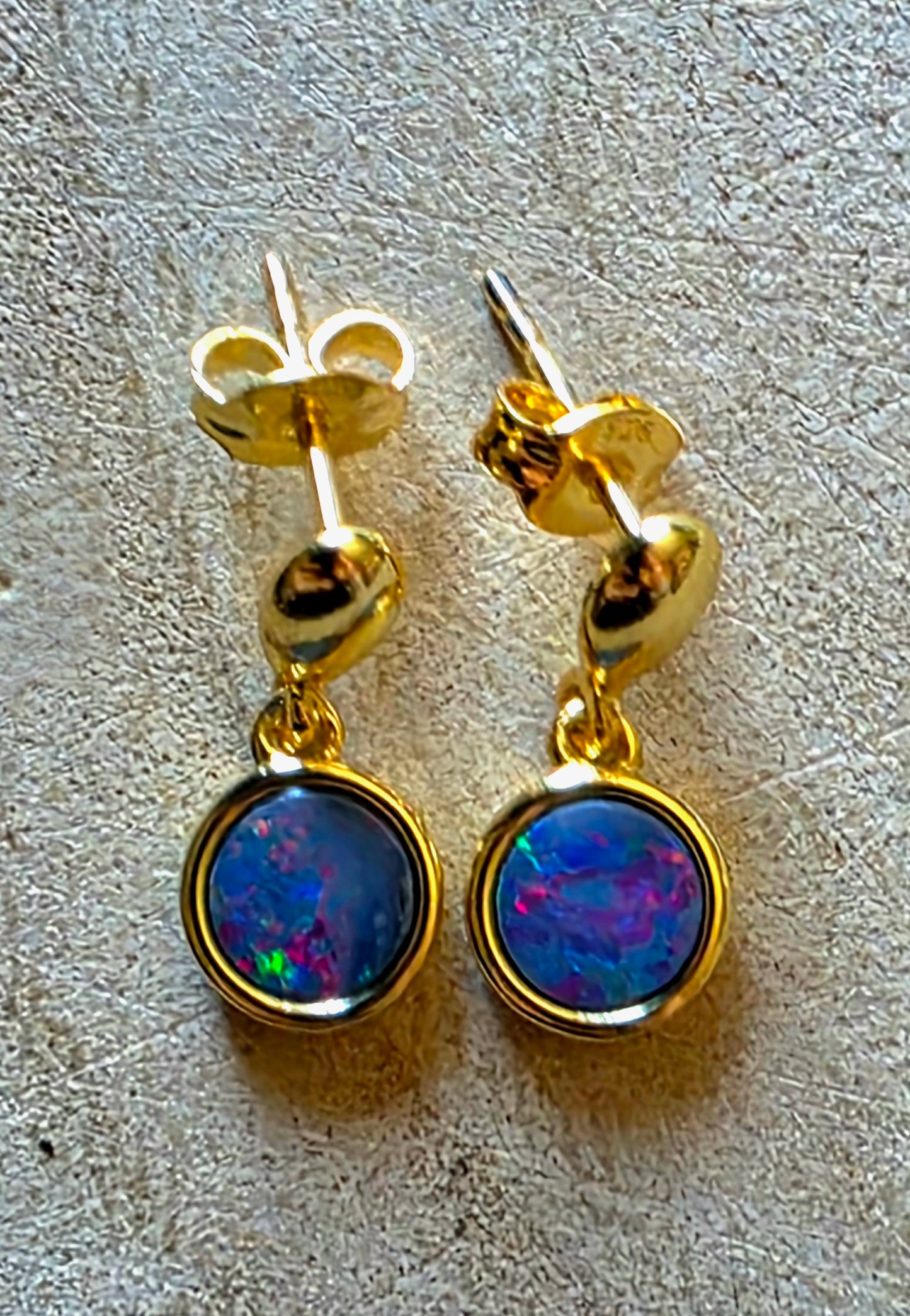 A pair of gold drop earrings with round bezel-set iridescent opal stones, displayed on a textured light beige surface.