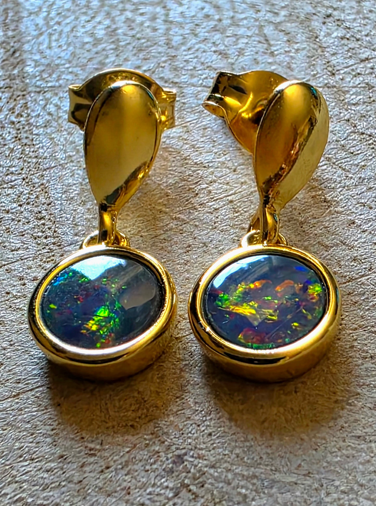 A pair of gold drop earrings with oval bezel-set iridescent opal stones, displayed on a textured beige surface.