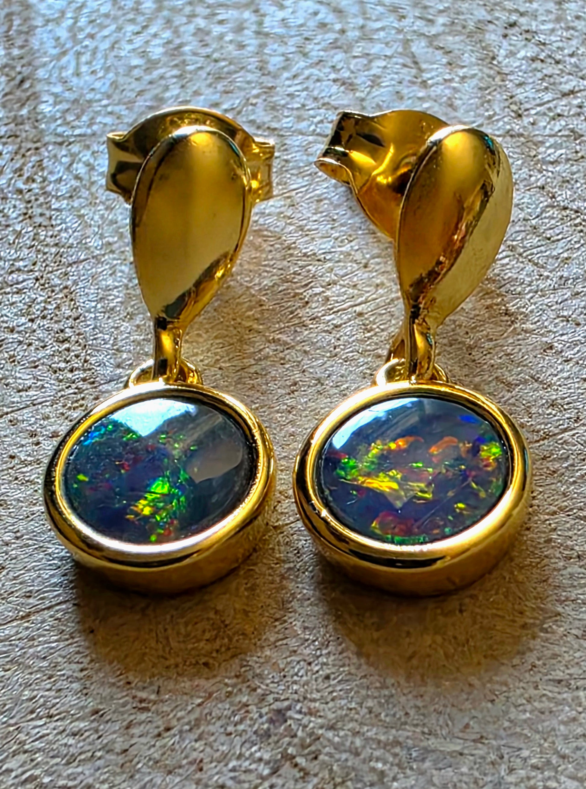 A pair of gold drop earrings with oval bezel-set iridescent opal stones, displayed on a textured beige surface.