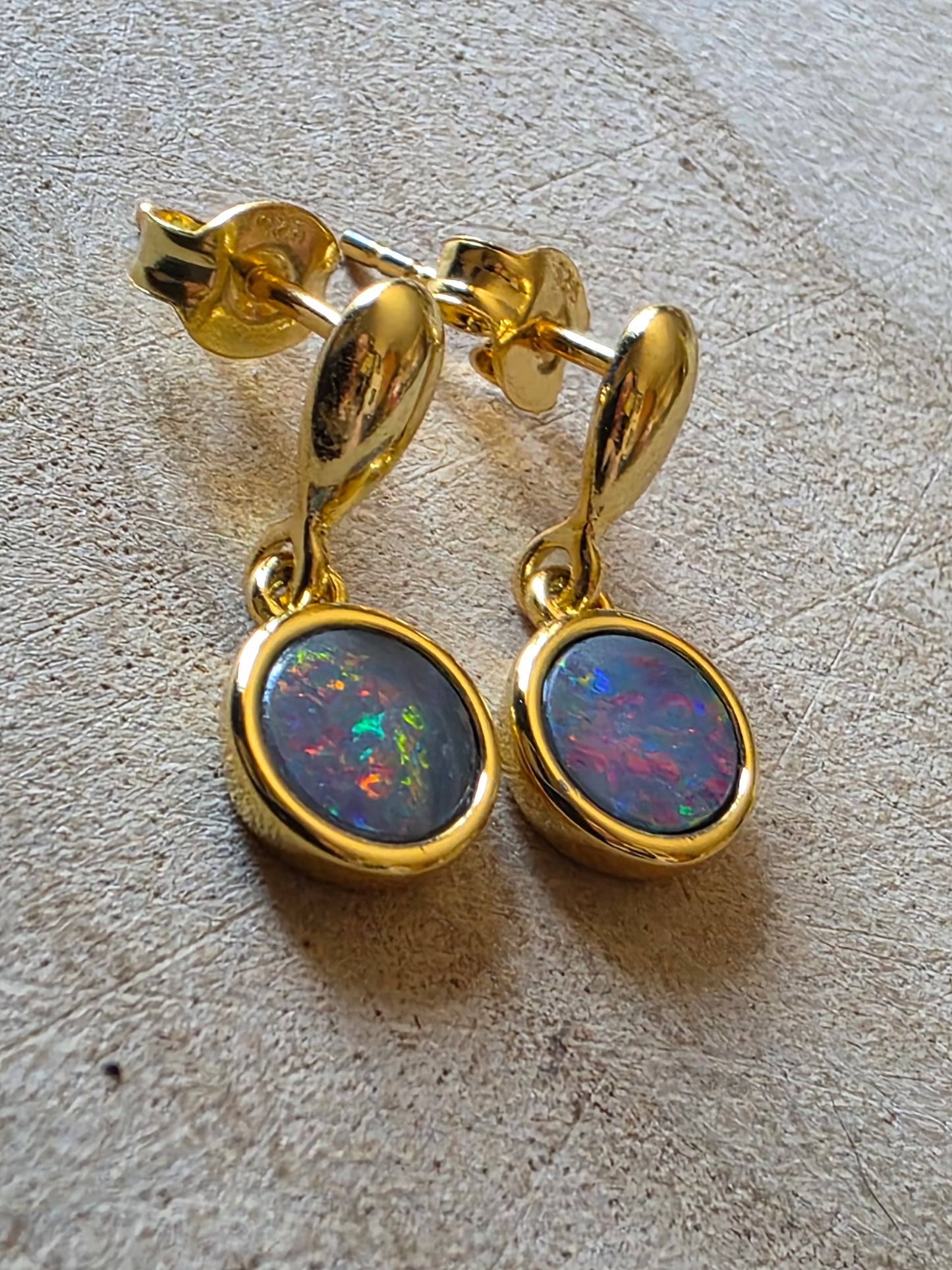 A pair of gold drop earrings with round bezel-set iridescent opal stones, displayed on a textured light surface.