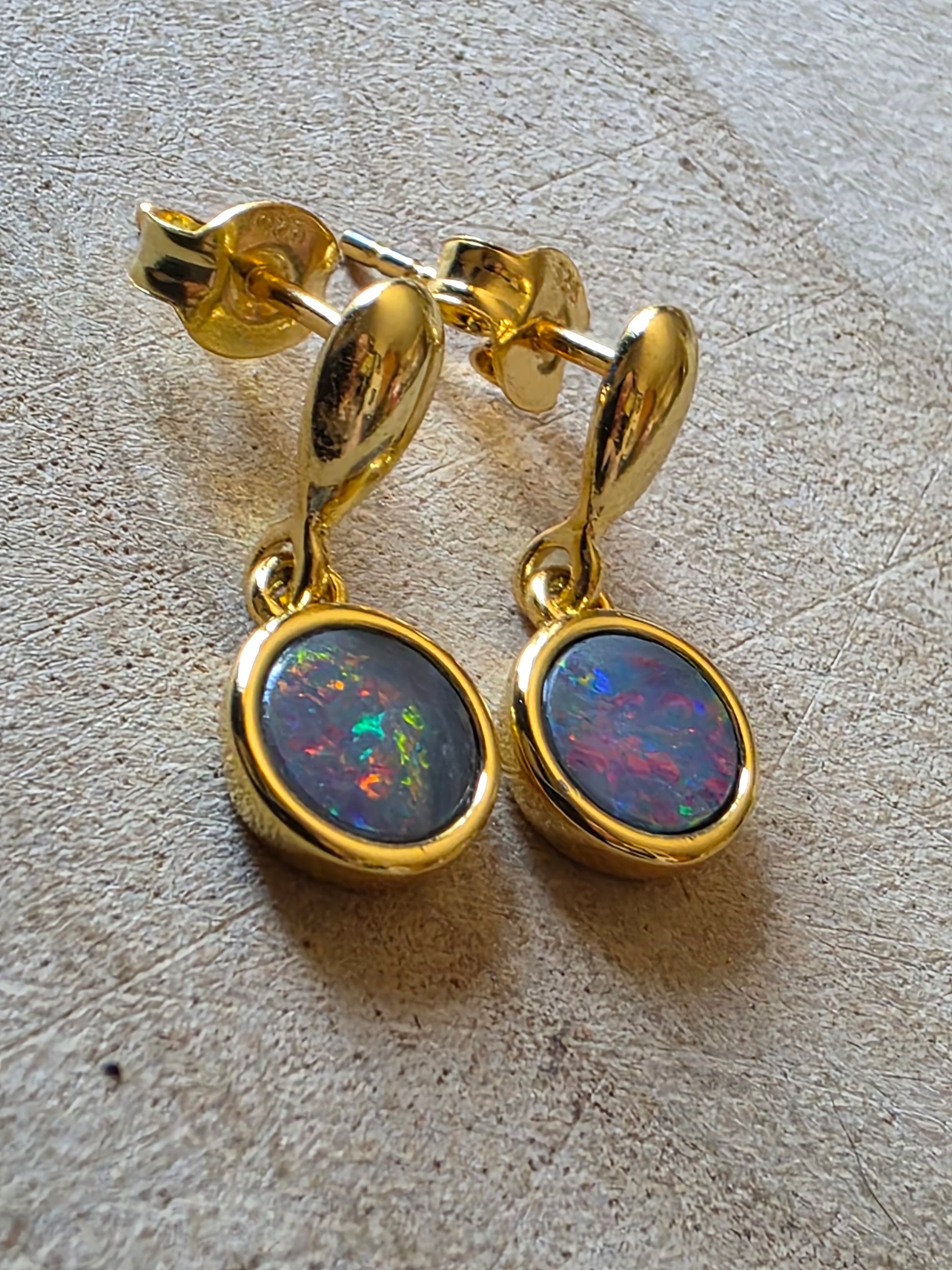 A pair of gold drop earrings with round bezel-set iridescent opal stones, displayed on a textured light surface.