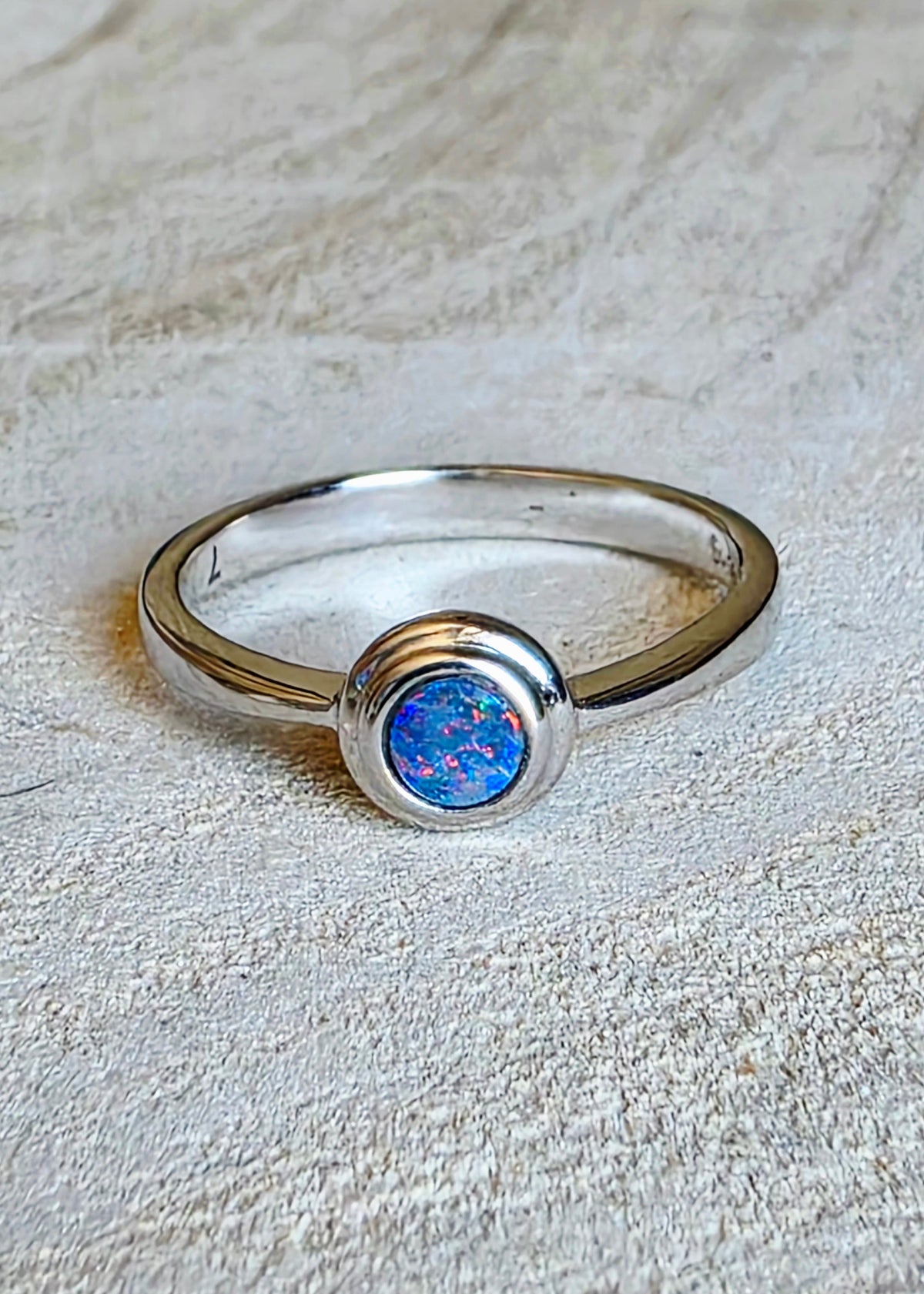A silver ring featuring a round bezel-set blue opal stone, displayed on a textured beige fabric background.