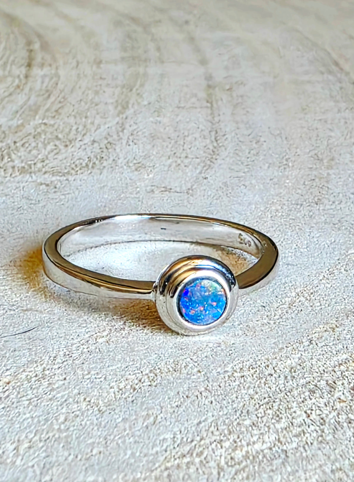 A silver ring with a round bezel-set blue opal stone, resting on a textured beige surface.