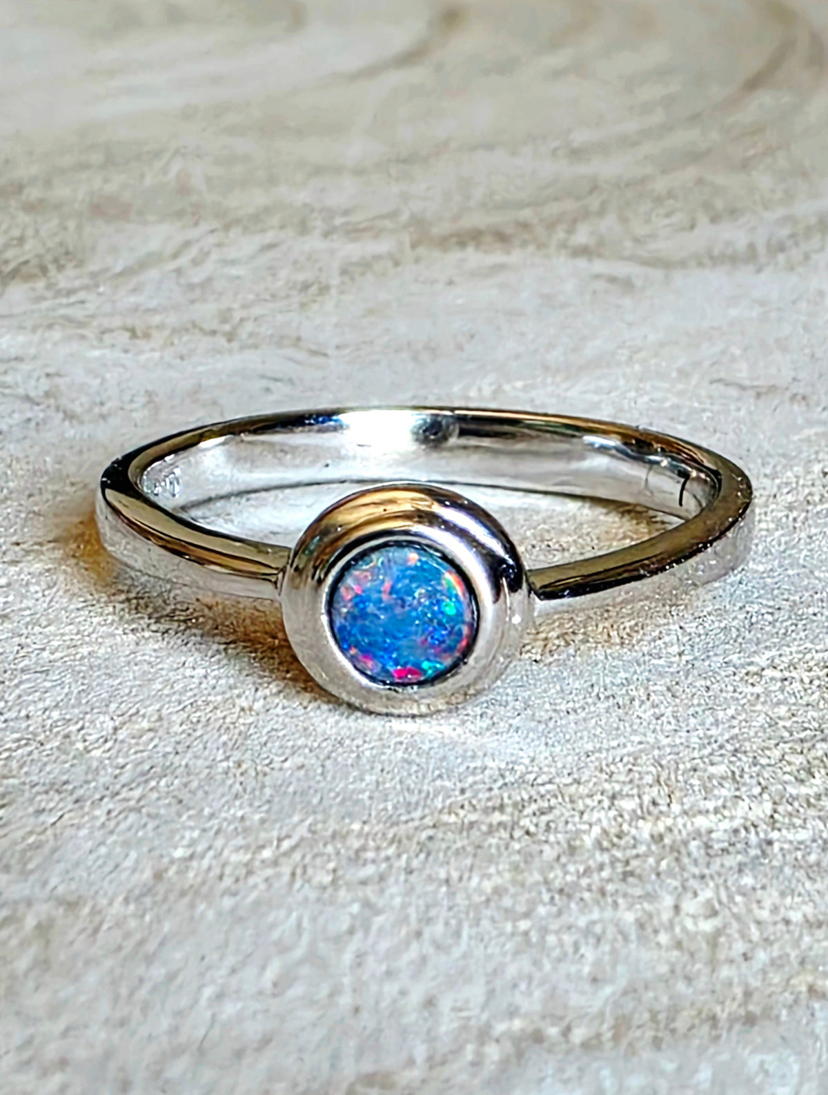 A sterling silver ring featuring a round bezel-set iridescent opal gemstone with vibrant blue, pink, and green flashes, resting on a textured beige fabric background.
