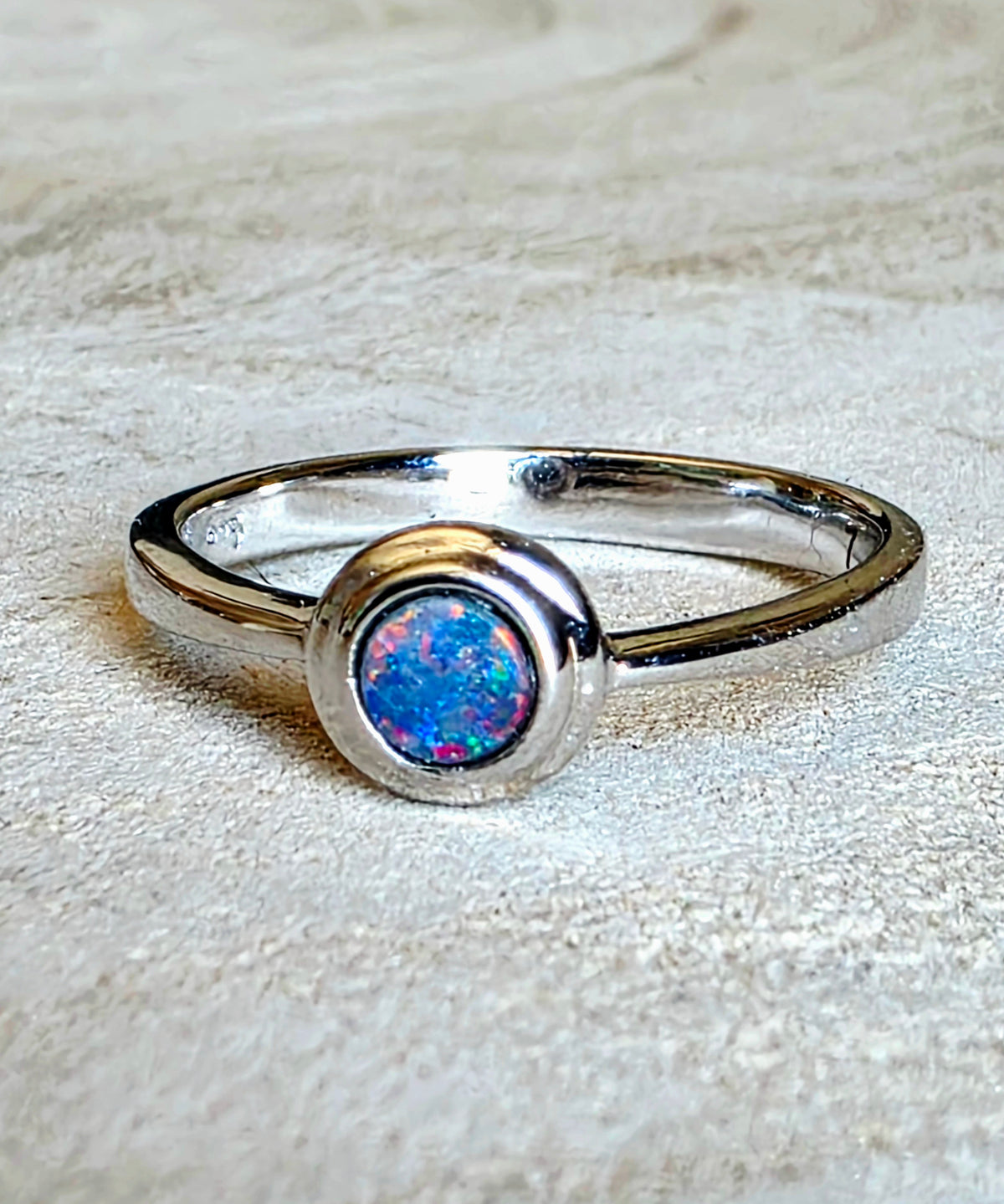 A silver ring featuring a round bezel-set iridescent opal stone with vibrant blue, pink, and green flashes, resting on a textured beige background.