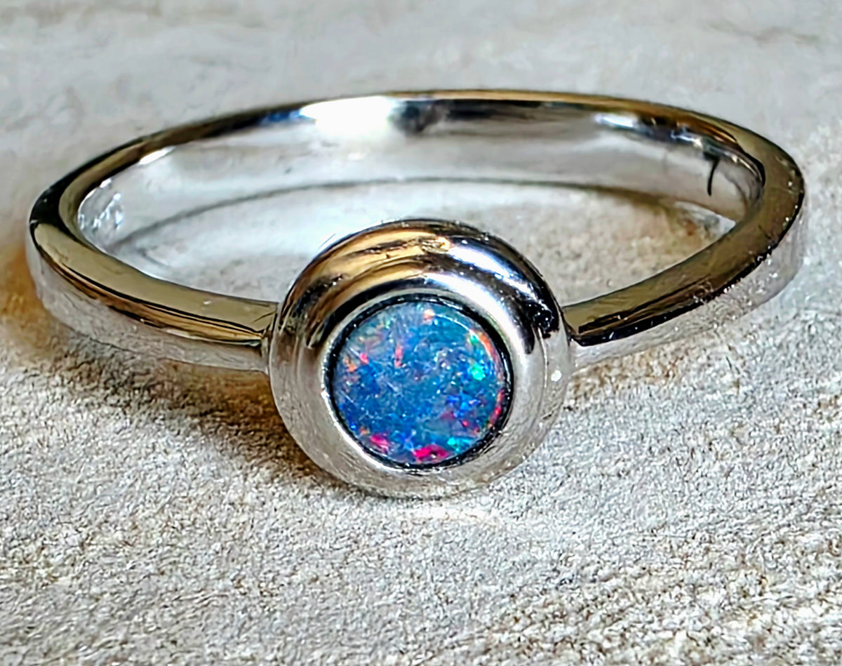 A polished sterling silver ring featuring a round bezel-set cabochon opal with vibrant blue, pink, and green flashes, resting on a textured light beige surface.