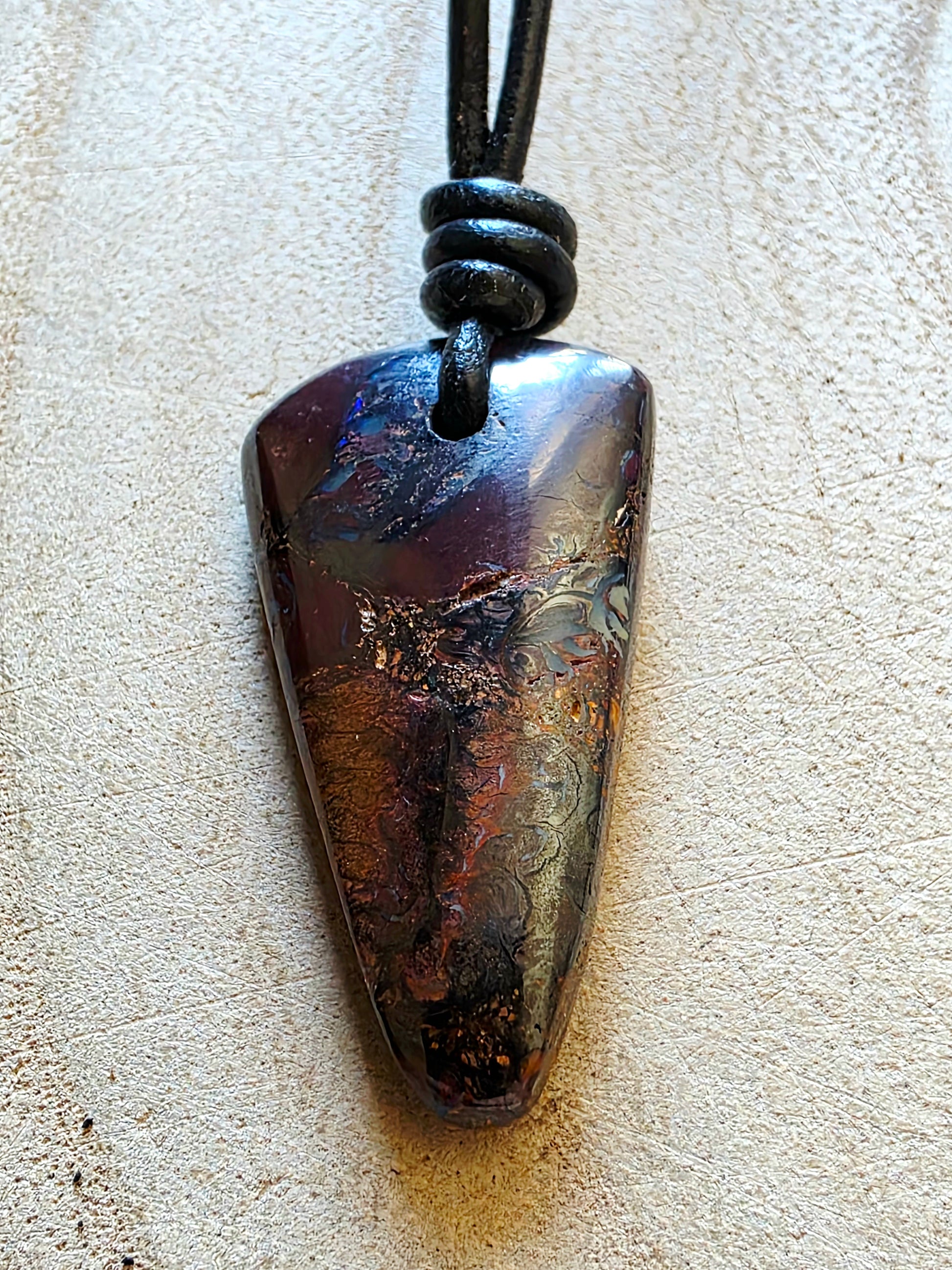 A polished triangle-shaped dark stone pendant with iridescent brown, purple, and gold marbling, suspended from a black cord on a textured beige surface.