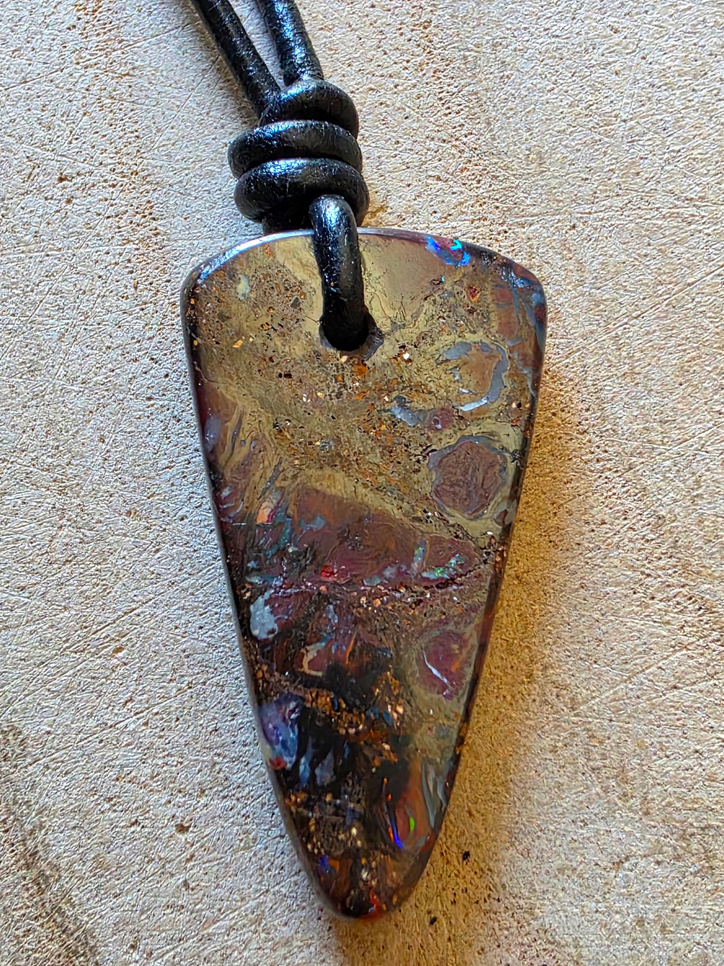 A natural opal pendant necklace with a triangle-shaped stone on a black cord, displayed on a textured beige surface.
